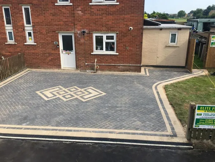 New block paving driveway installation with grey blocks, a beige border, and a decorative pattern, featuring an Aco drain, by Elite Paving and Roofing LTD.