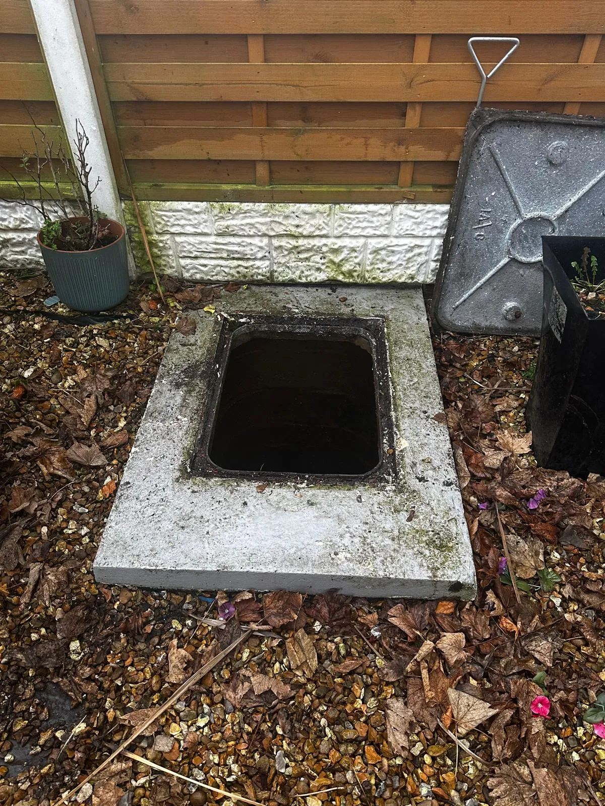 Open concrete manhole access point in a residential garden, with the cover lifted and resting against a wooden fence, surrounded by gravel and autumn leaves.