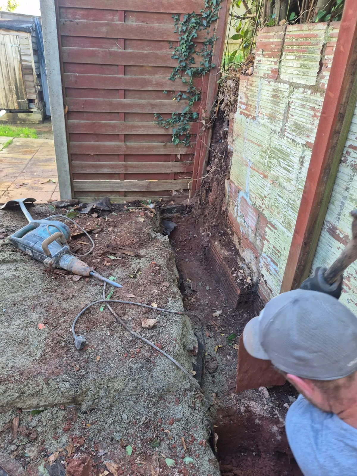 A construction worker carrying out groundworks, digging a trench next to a fence and an old wall, with a jackhammer lying nearby. The ground is excavated earth and rubble.