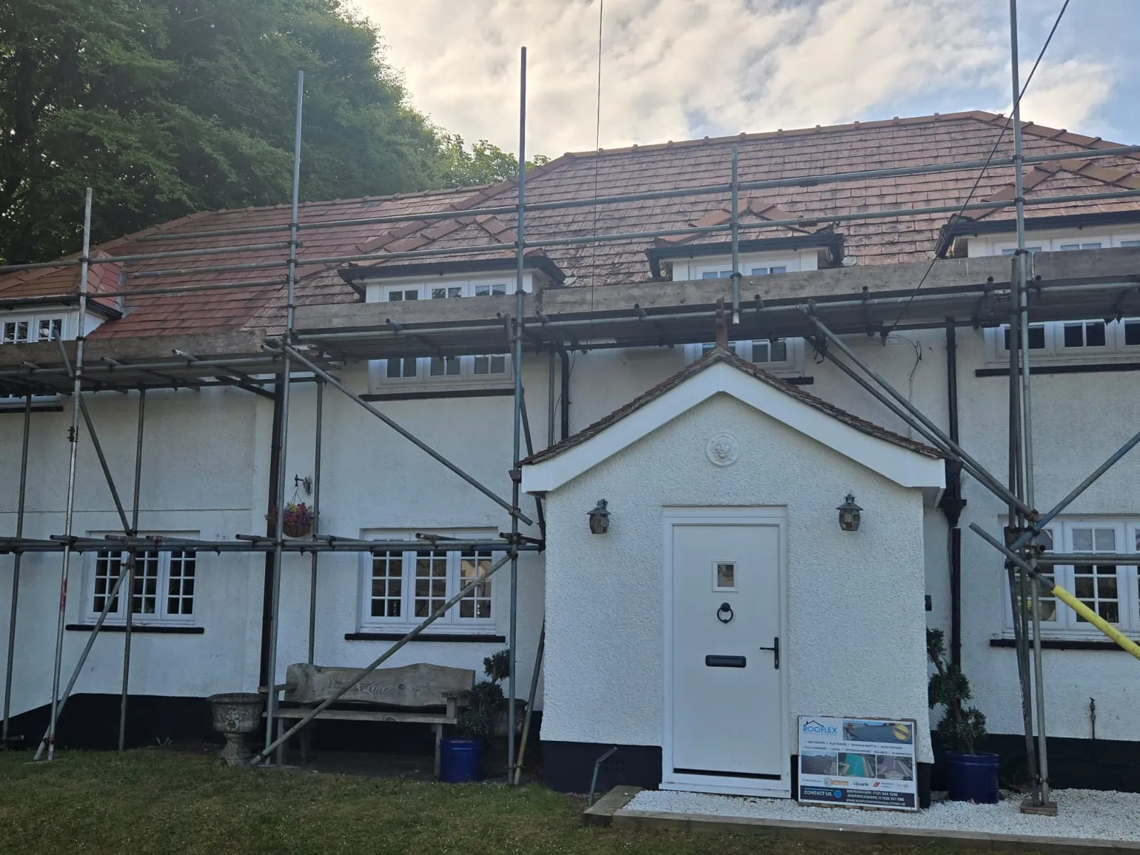 A house with scaffolding erected for a re-roofing project, showing a partially stripped red tiled roof and a new white front door.