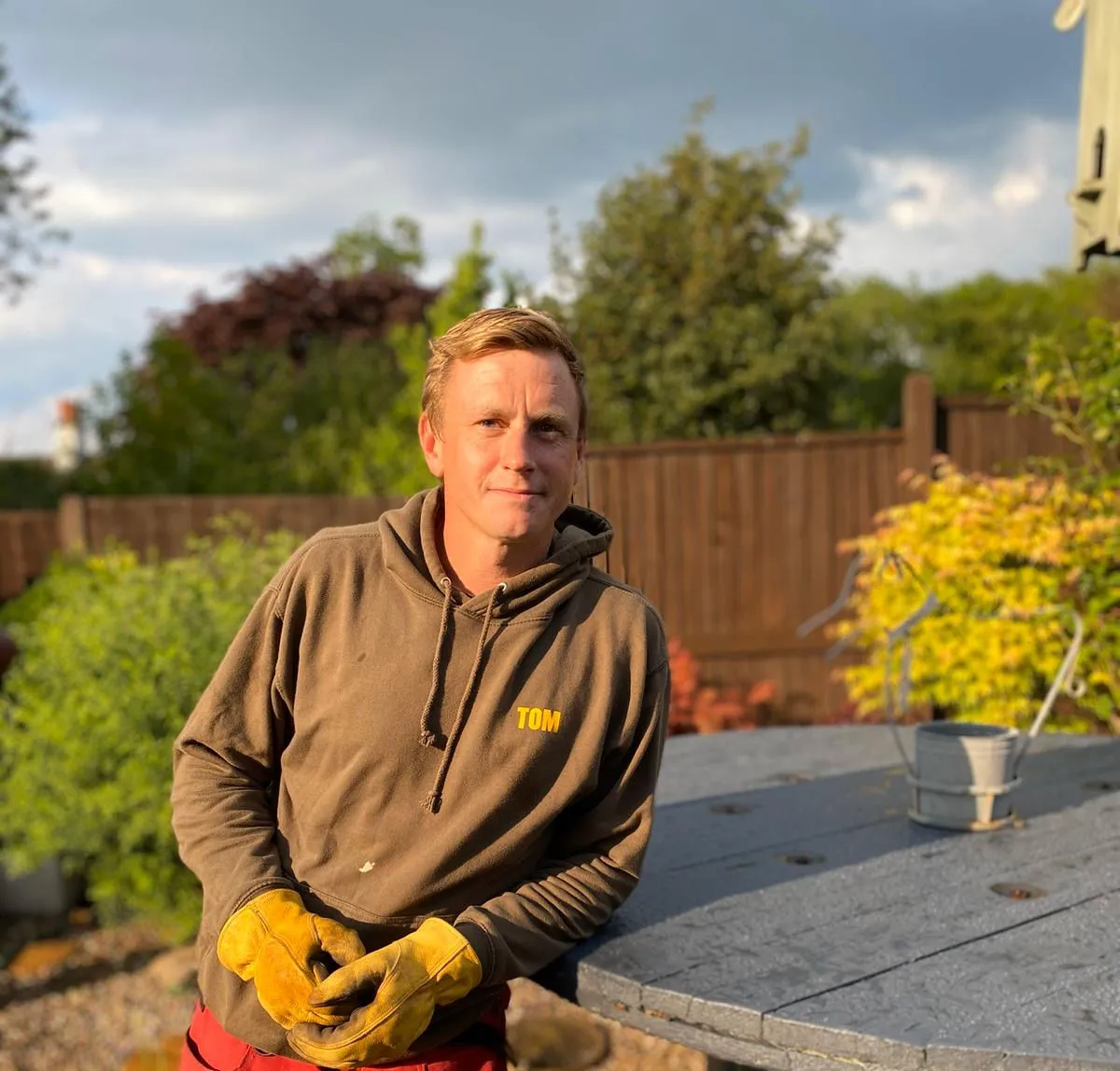 A man in a brown work hoodie with 'TOM' on it and yellow gloves, smiling in a garden.