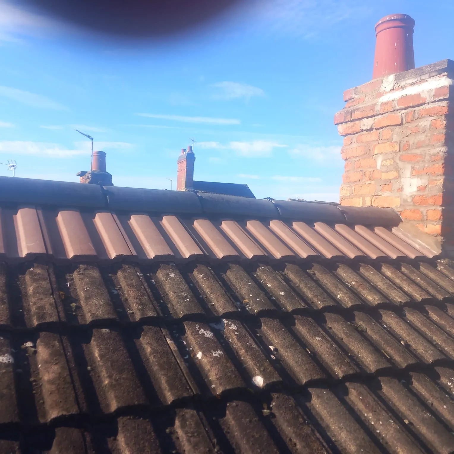 Close-up of a tiled roof showing a mix of old, mossy tiles and cleaner, newer tiles, with brick chimneys against a blue sky.