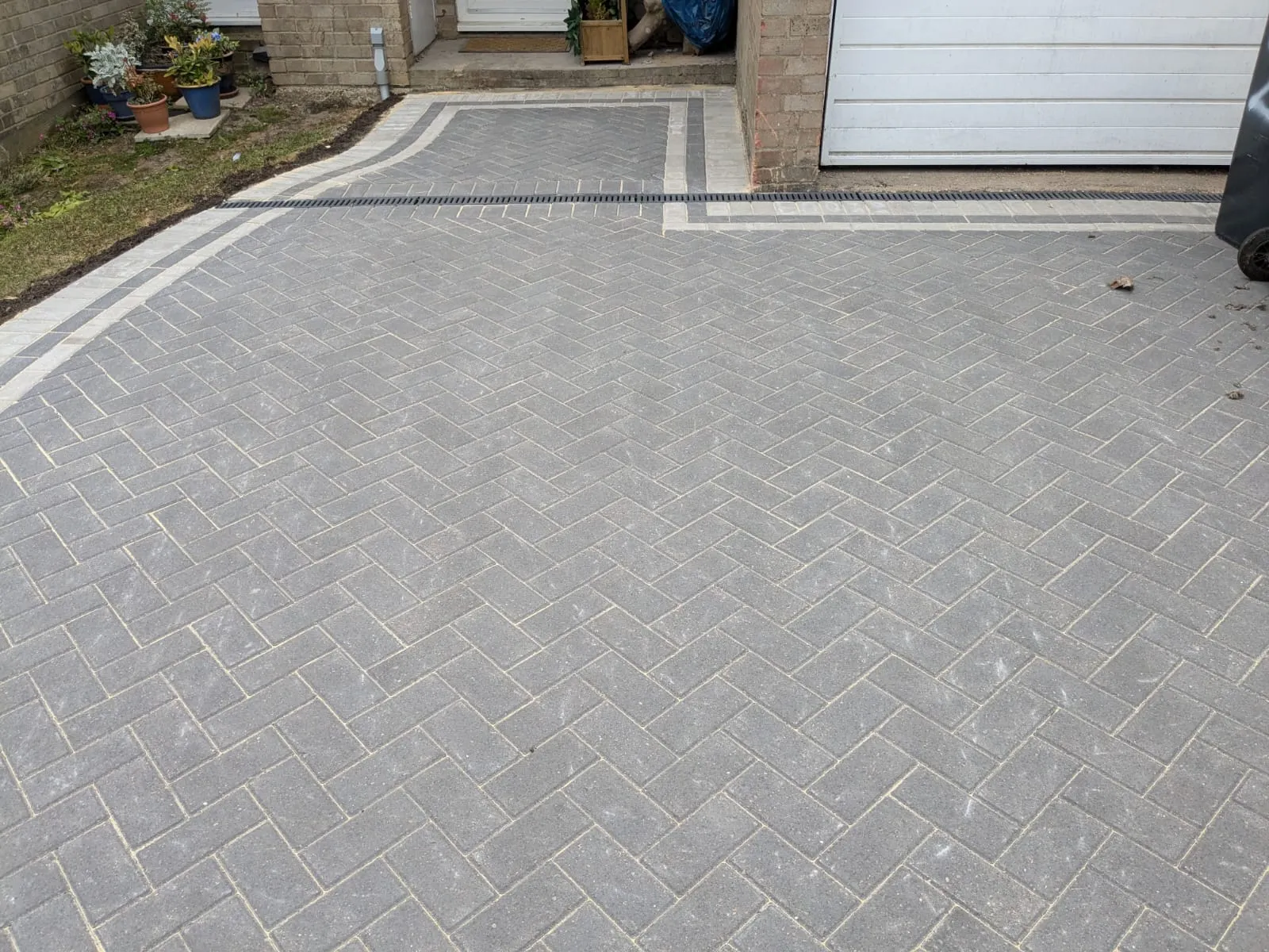 A modern grey block paved driveway with a herringbone pattern, light border, and drainage system, leading to a house and garage.