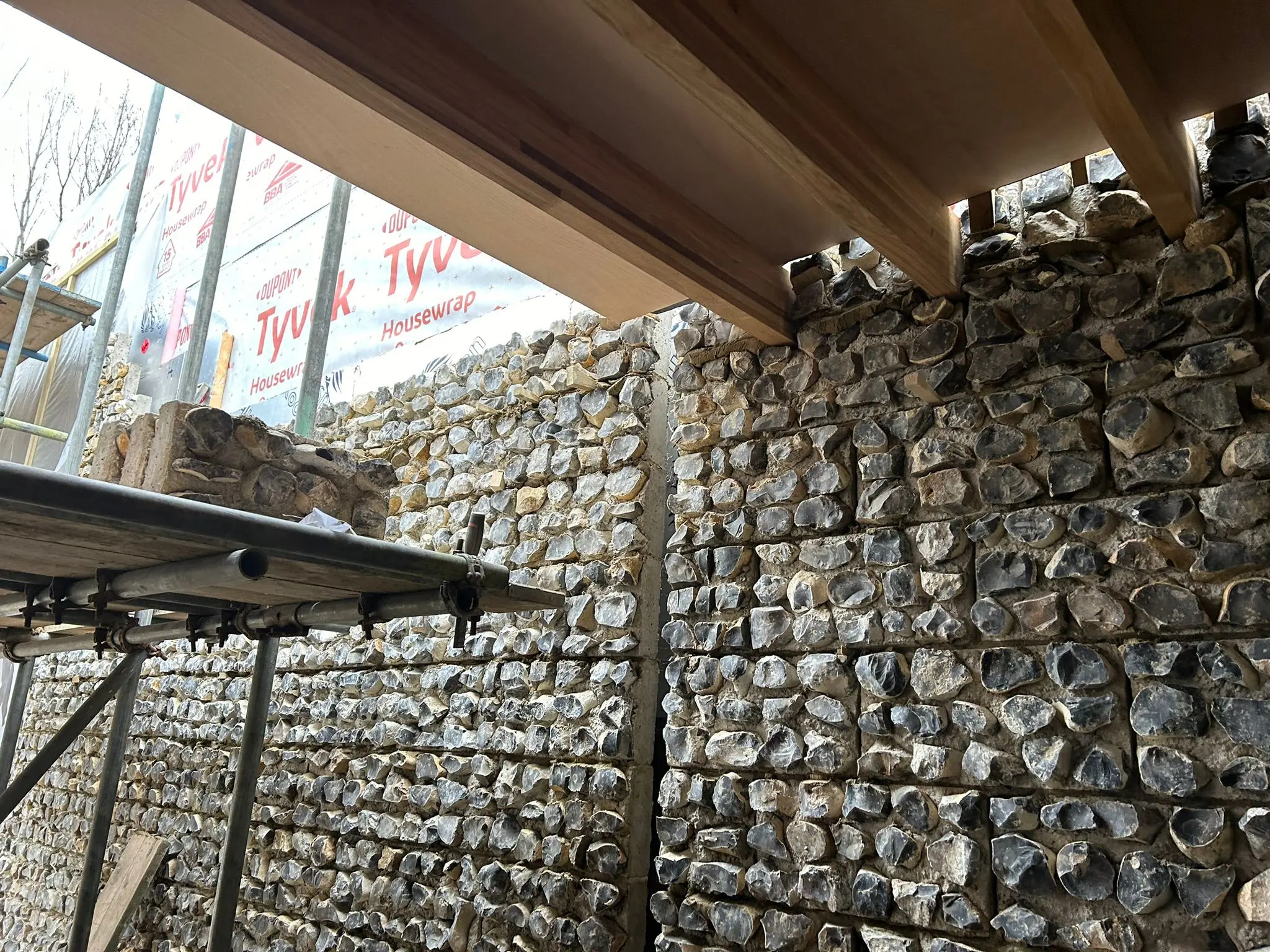 A close-up shot of a flint wall being constructed with scaffolding and house wrap visible.