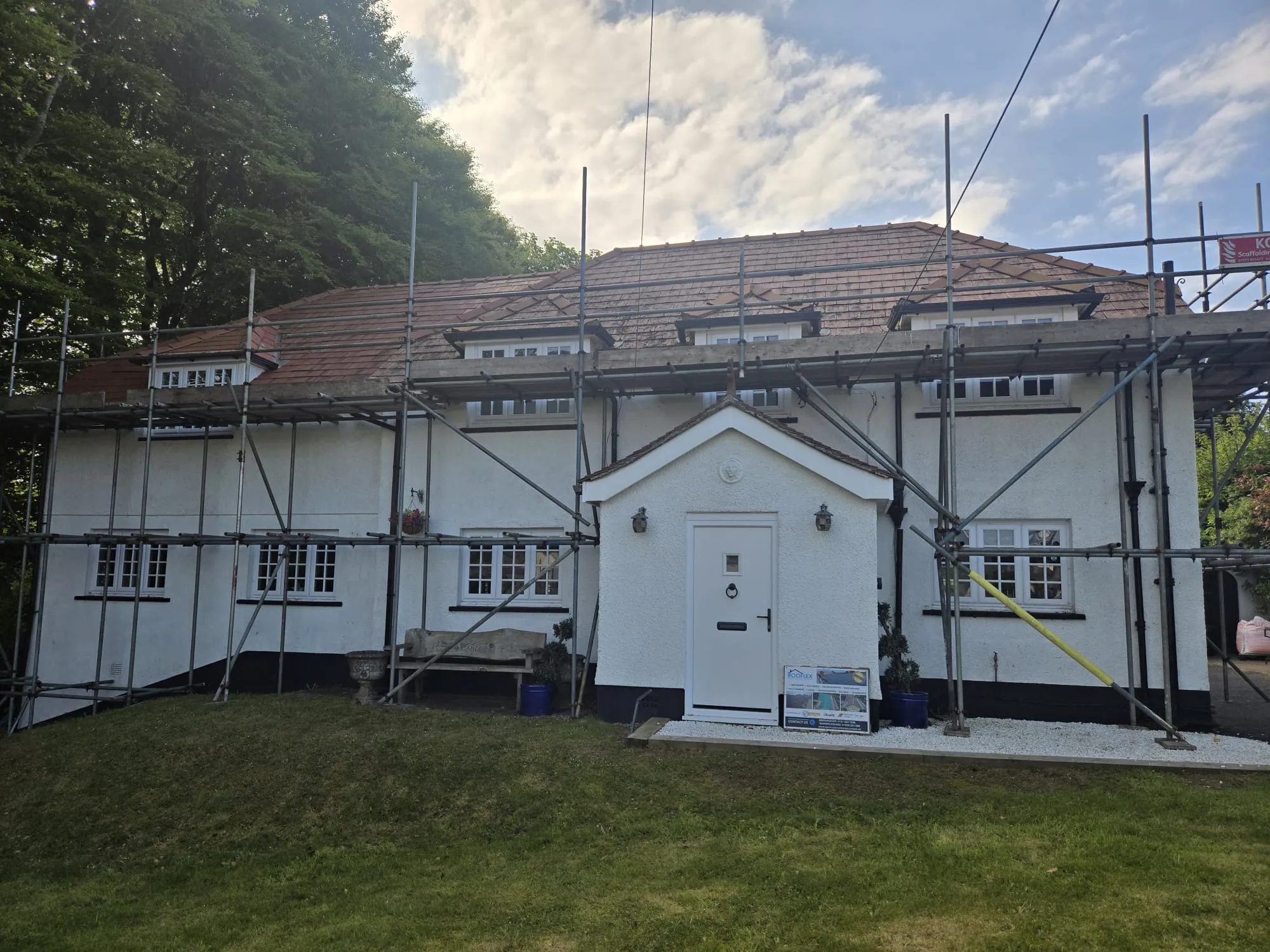 A white residential house with a new red tiled roof, surrounded by scaffolding during a re-roofing project, with a Rooflex Roofing Solutions sign visible.