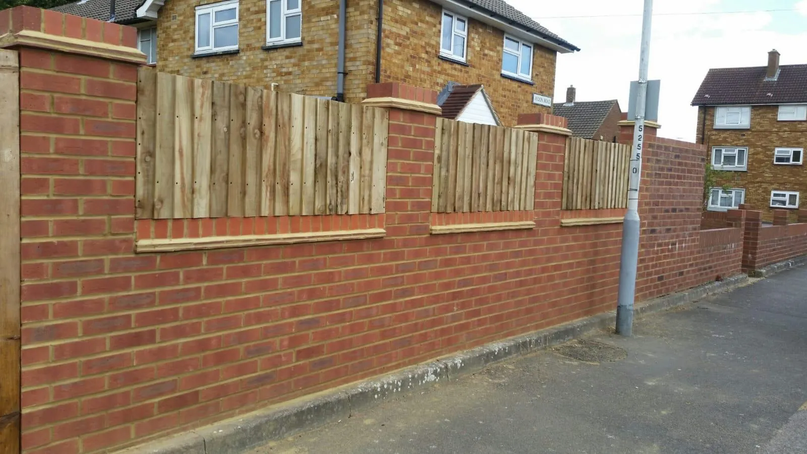 A new red brick garden wall with wooden fence panels and decorative brick caps, bordering a pavement with houses in the distance.