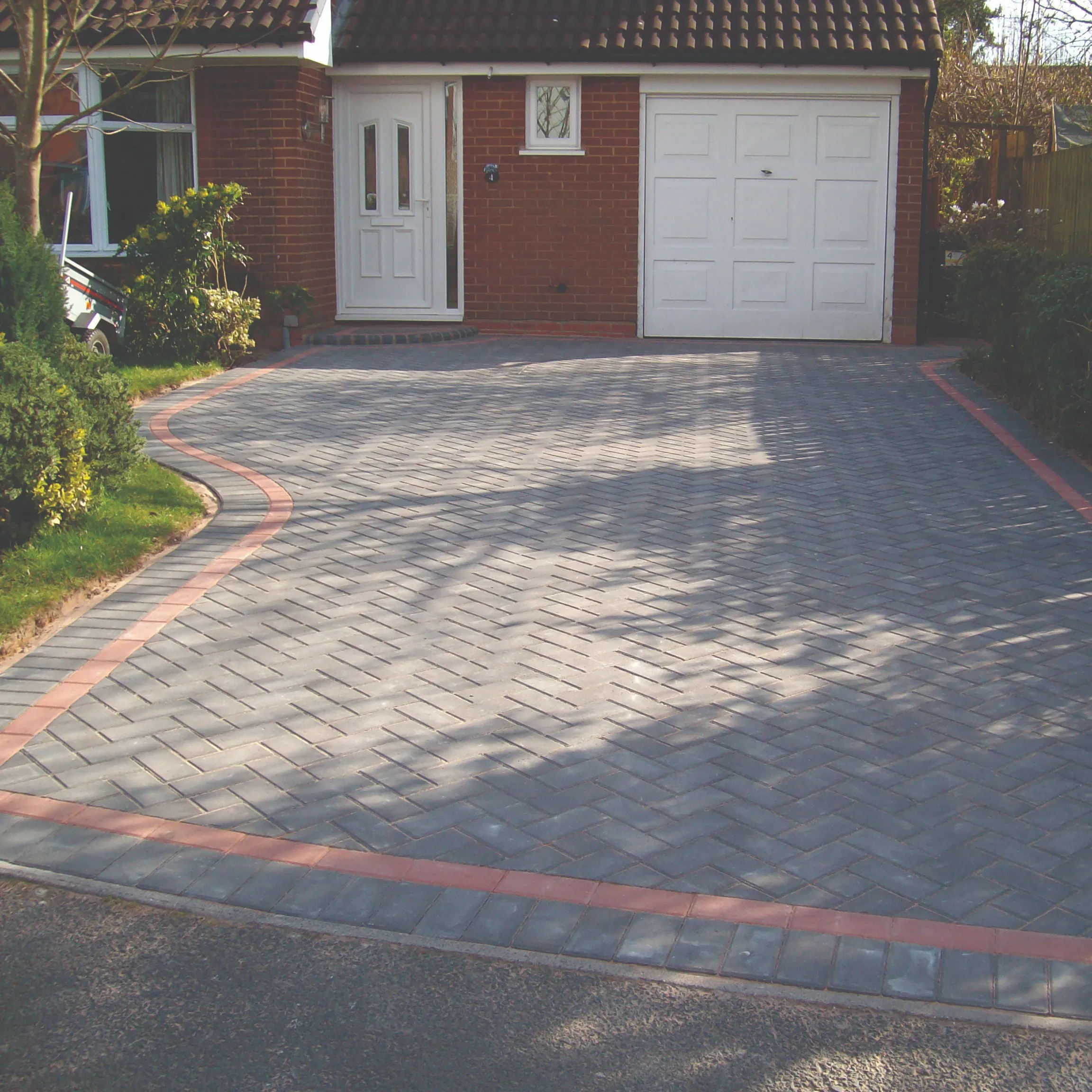 A modern grey block paving driveway with a red block border, featuring a curved edge next to a lawn, leading to a red brick house with a white front door and garage door.
