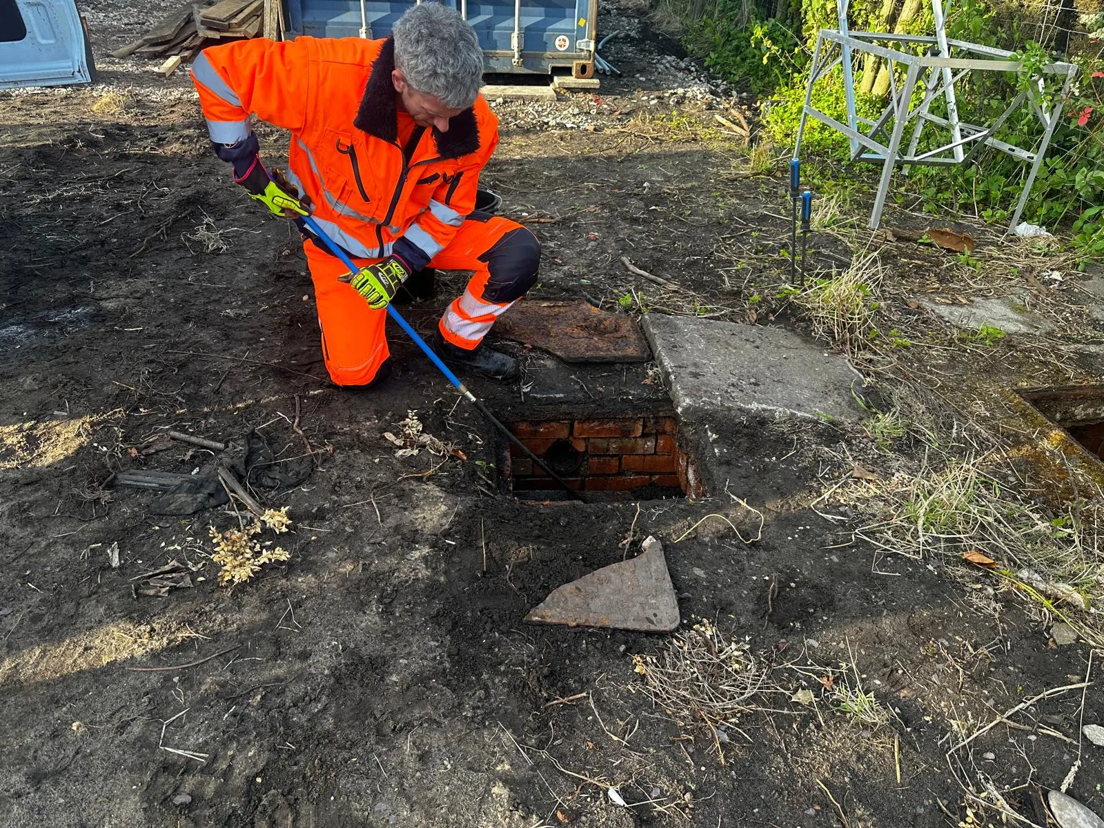 Drainage technician in a high-vis suit using a drain rod to clear an external manhole.