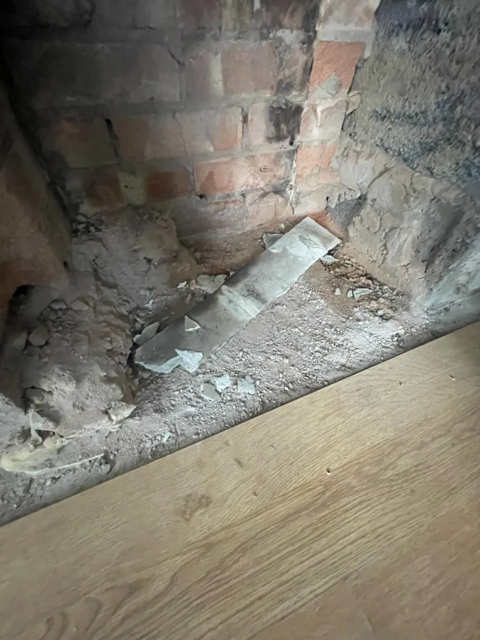 Broken suspected asbestos material on disturbed ground next to a brick wall and wooden floor.