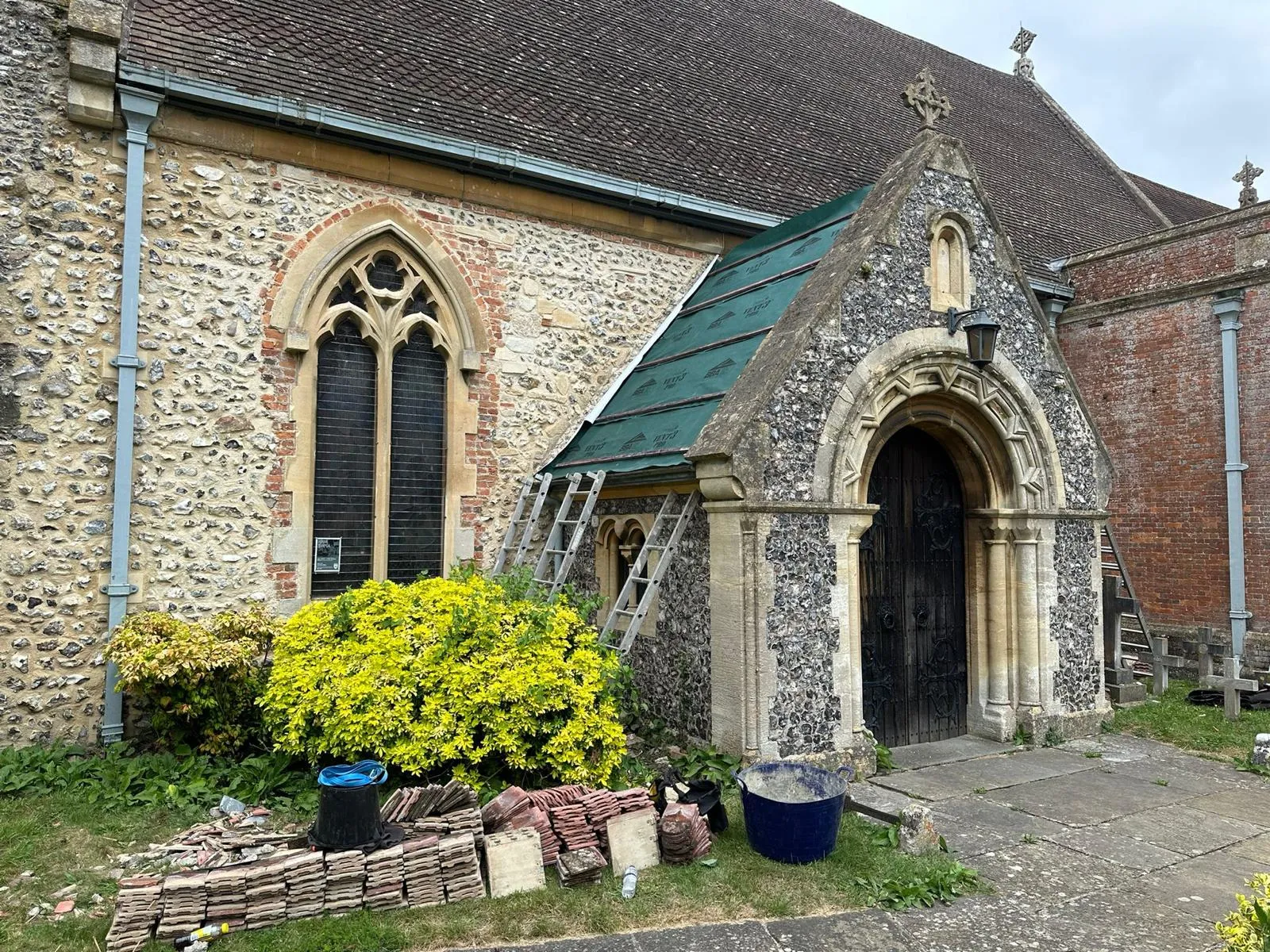 Work in progress on a church roof, with new green underlay installed on a section of the roof and stacks of old terracotta and stone tiles on the grass ready for removal.