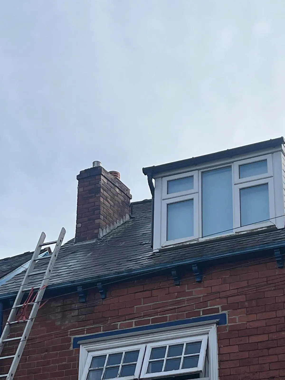 A slate roof with a brick chimney stack, uPVC dormer window, and a ladder set up for roof maintenance