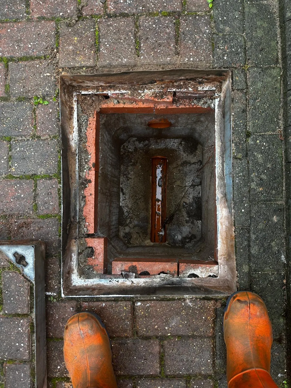 A picture looking down into an open drain manhole, showing standing water from a blockage, with a worker's boots in the foreground.