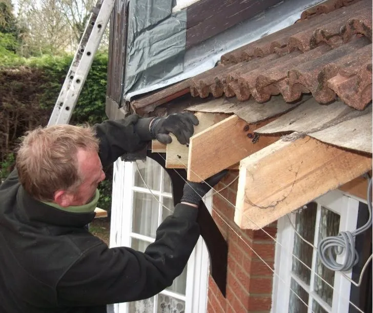 A roofer working on the timber elements of a roofline under existing tiles, with a ladder in the background.