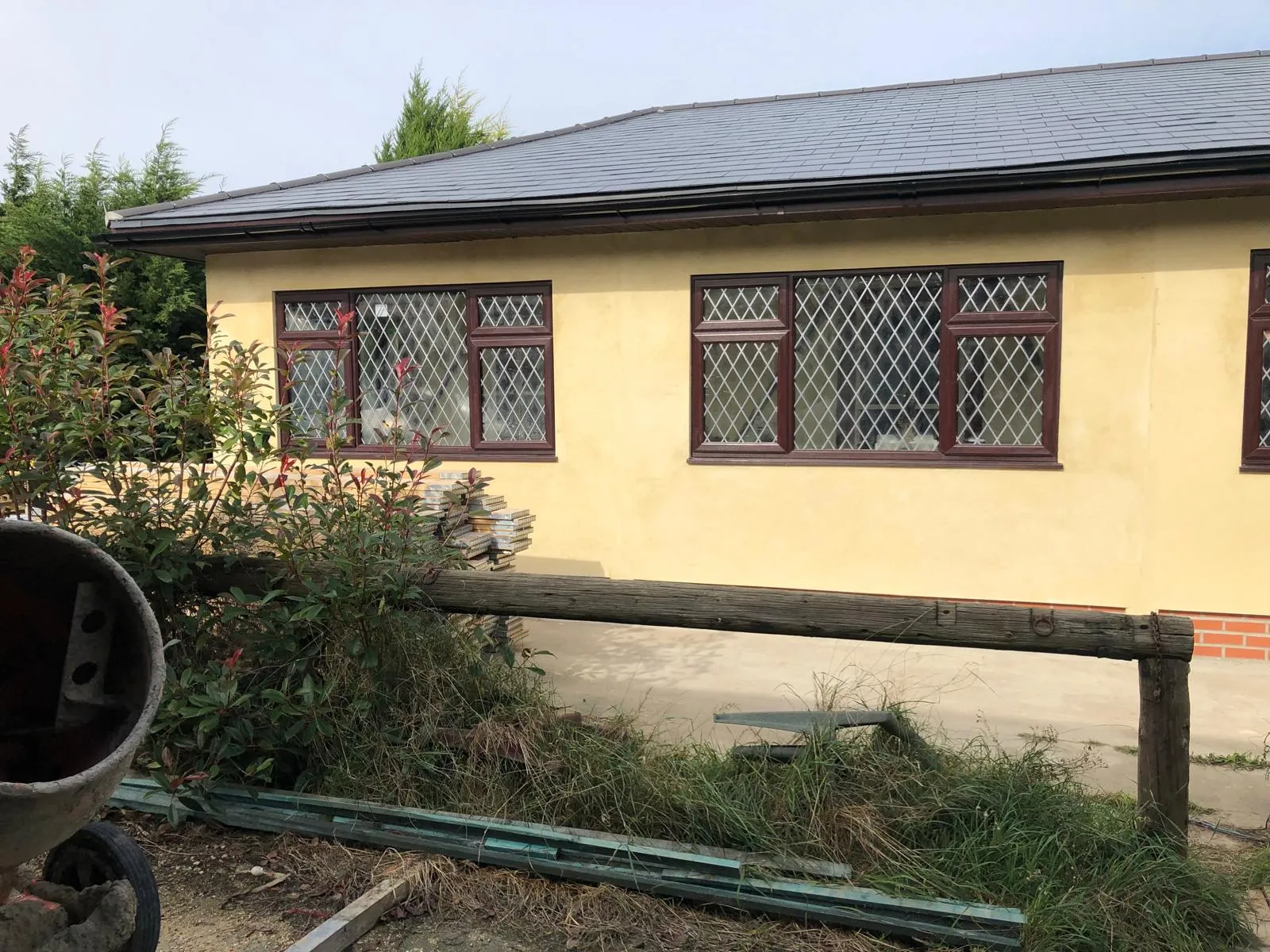 Exterior of a renovated house with yellow rendered walls, brown diamond-leaded windows, and a grey tiled roof, with a cement mixer and overgrown bushes in front.
