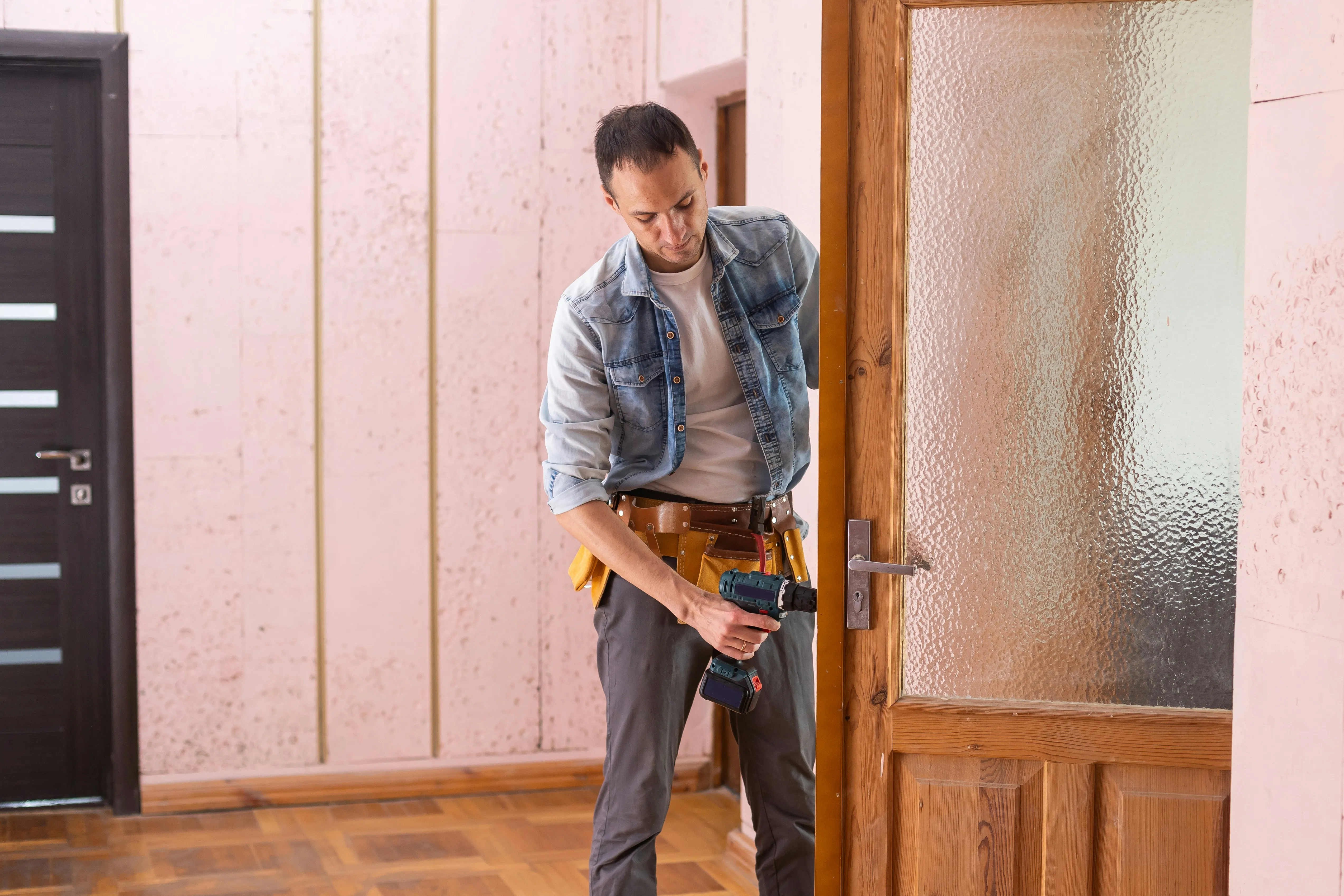 A locksmith wearing a tool belt, using a power drill to work on a lock on a wooden door with a frosted glass panel.