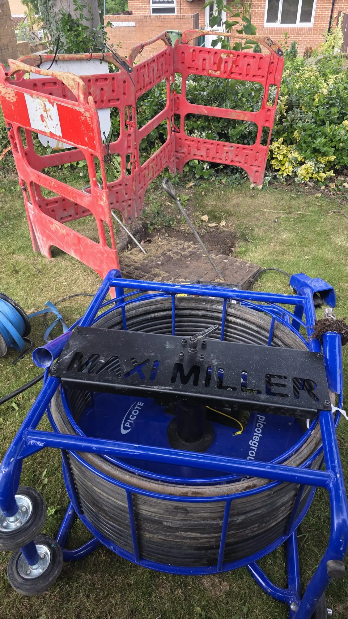 A blue Picote Maxi Miller drain milling machine set up on grass next to red safety barriers and an open manhole.