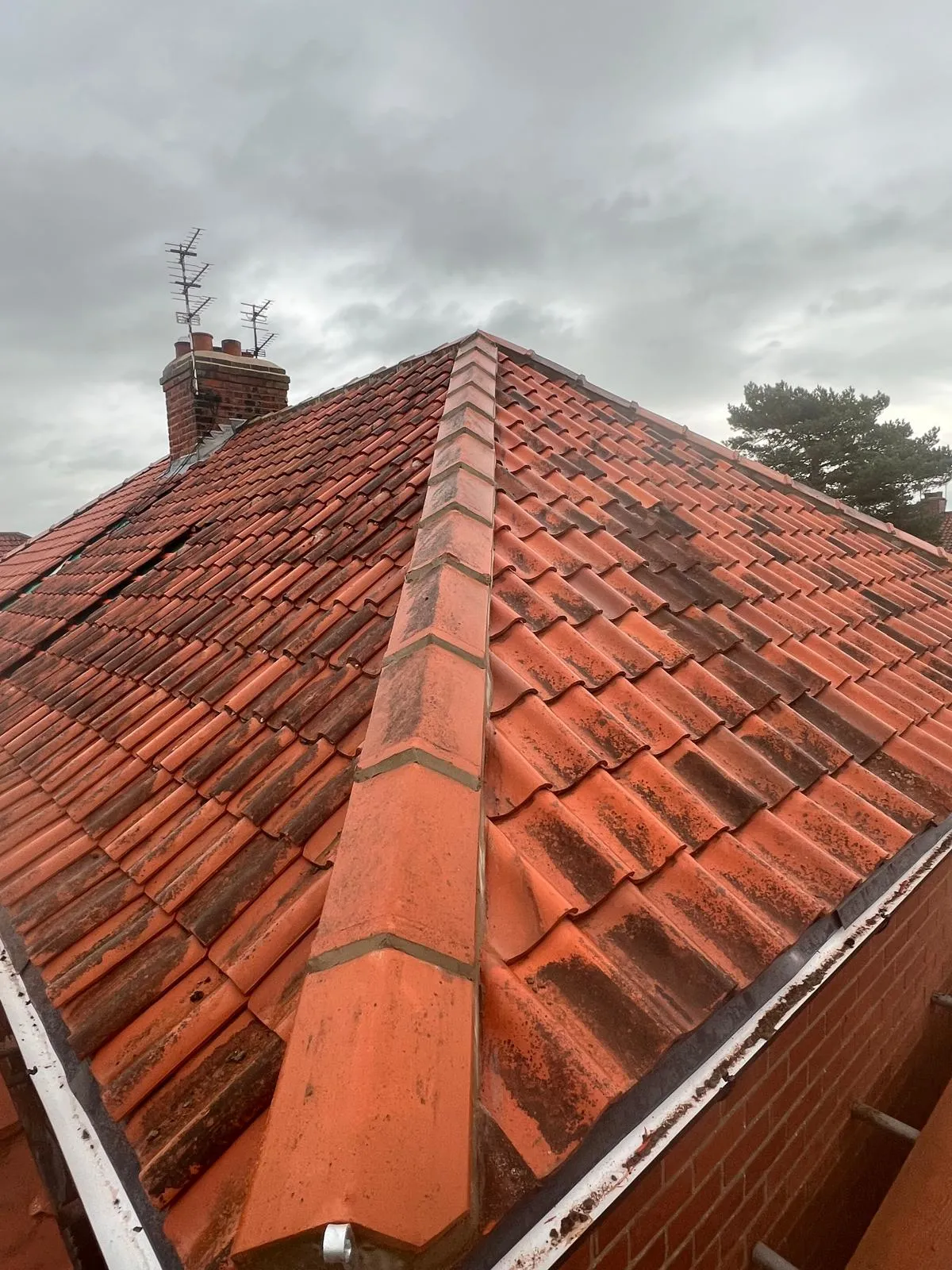 Weathered orange terracotta tiled pitched roof with a chimney stack and aerials, showing signs of wear and potential damage.