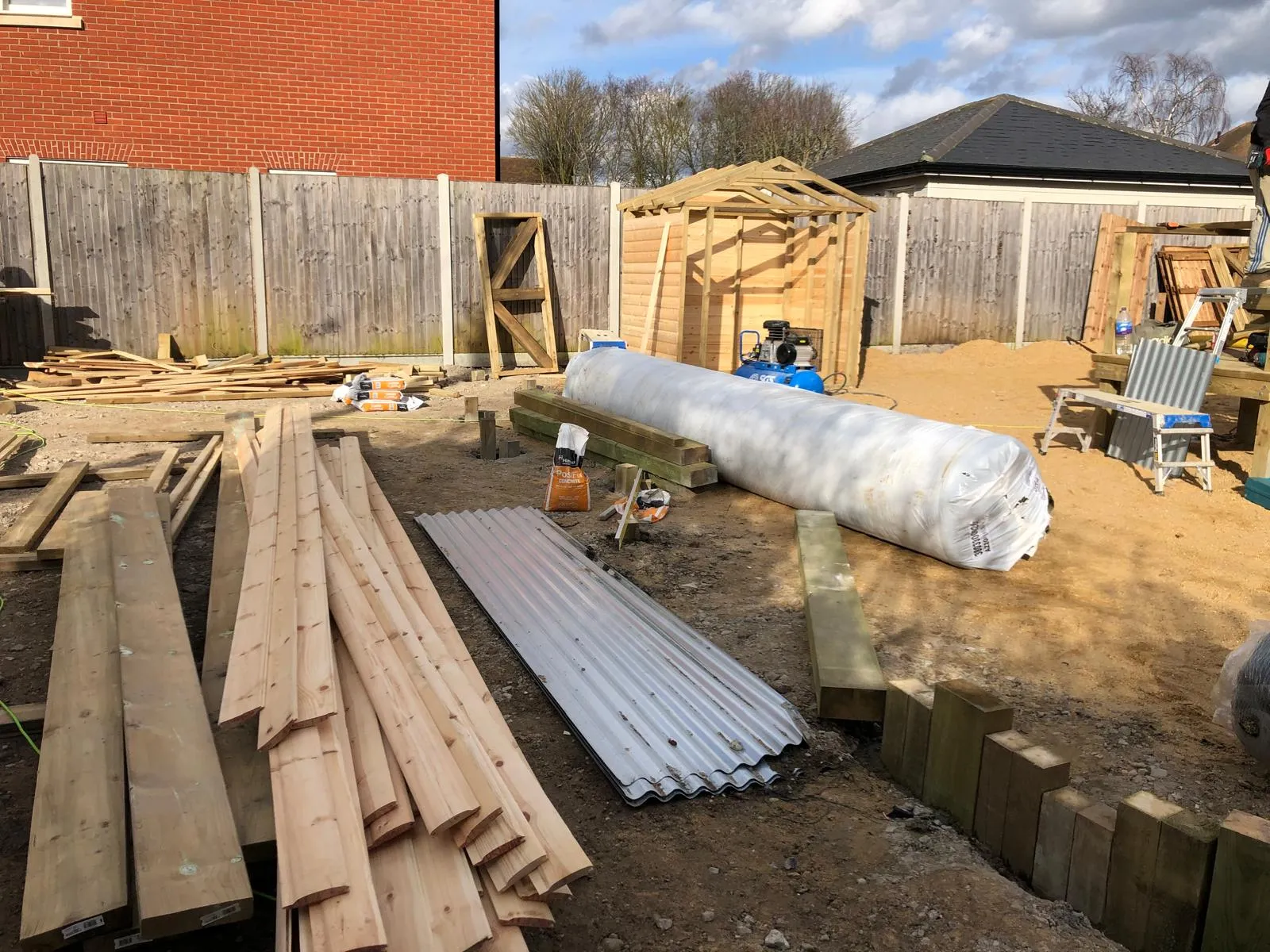 A construction site in a garden with building materials, including timber planks, corrugated metal, and the partially built wooden frame for a garden shed or office.