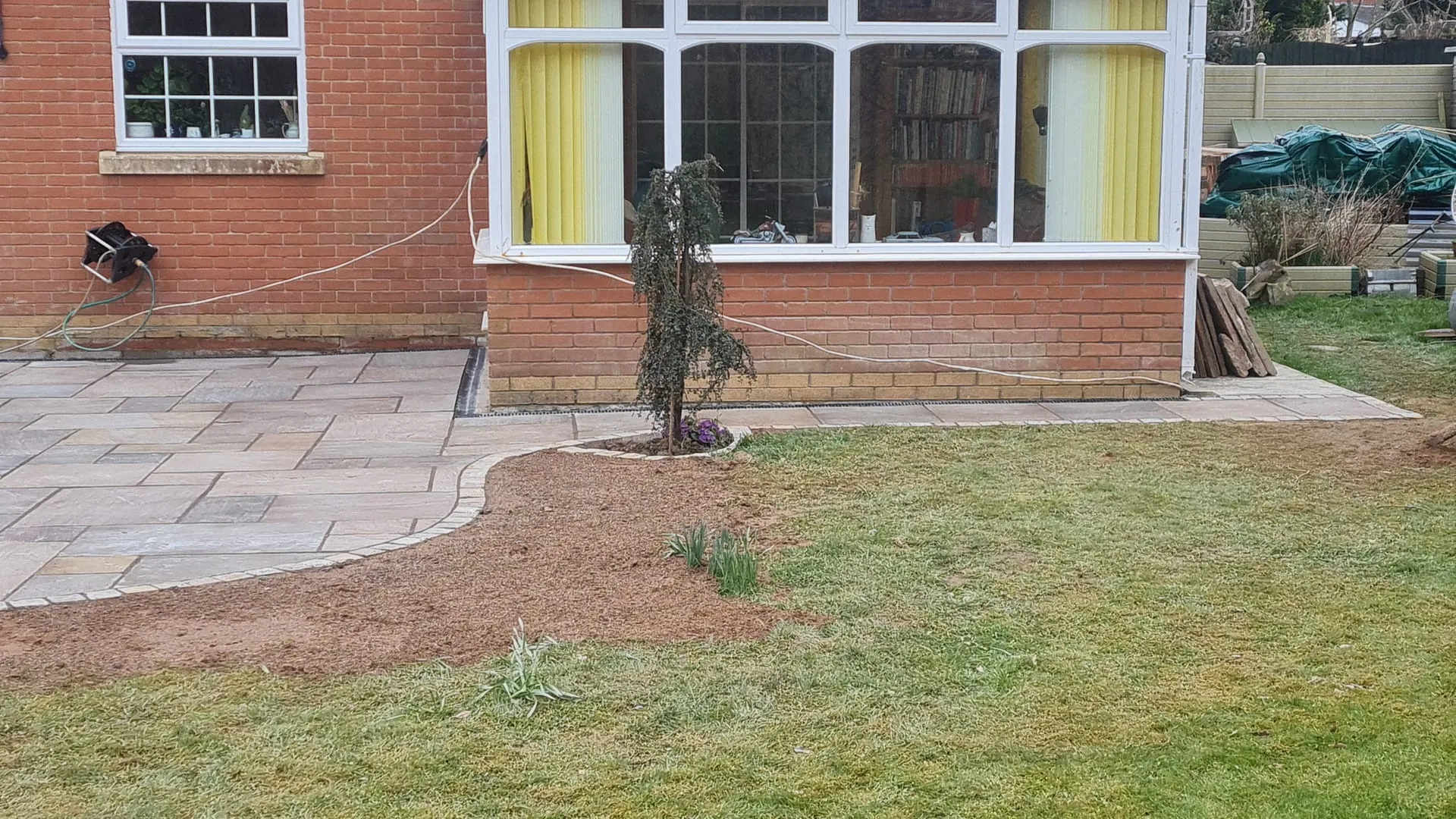 A professional new patio installation with light grey paving slabs, a decorative curved block paving edge, and a small tree planting next to a lawn and a house.