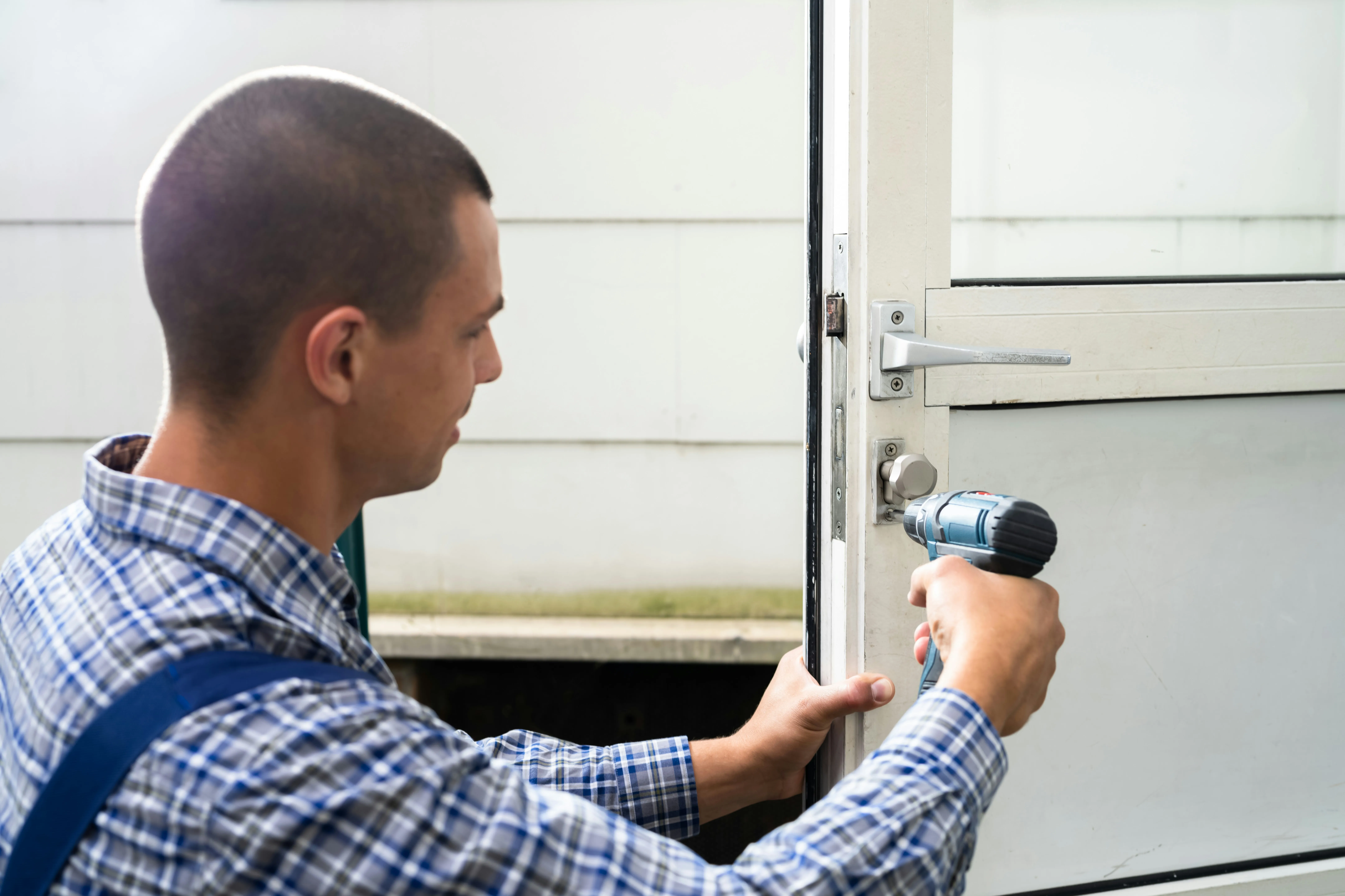 A close-up of a locksmith in a blue plaid shirt using a power drill to repair a uPVC door lock.