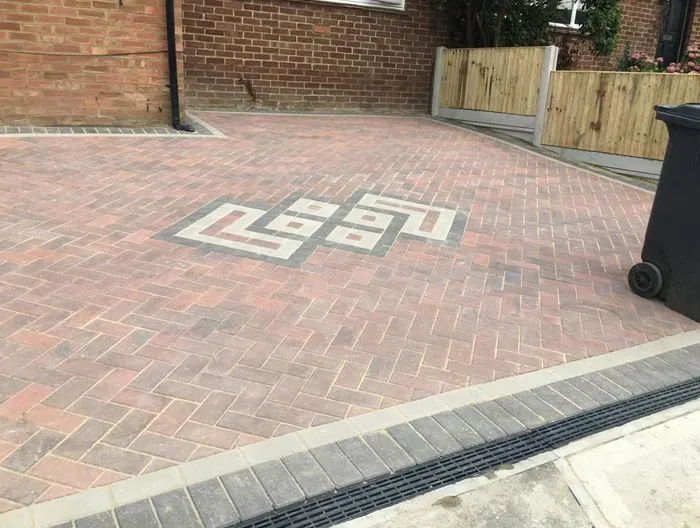 A completed block paving driveway with a decorative grey and light block pattern, bordered by a drainage channel and leading to a brick home.