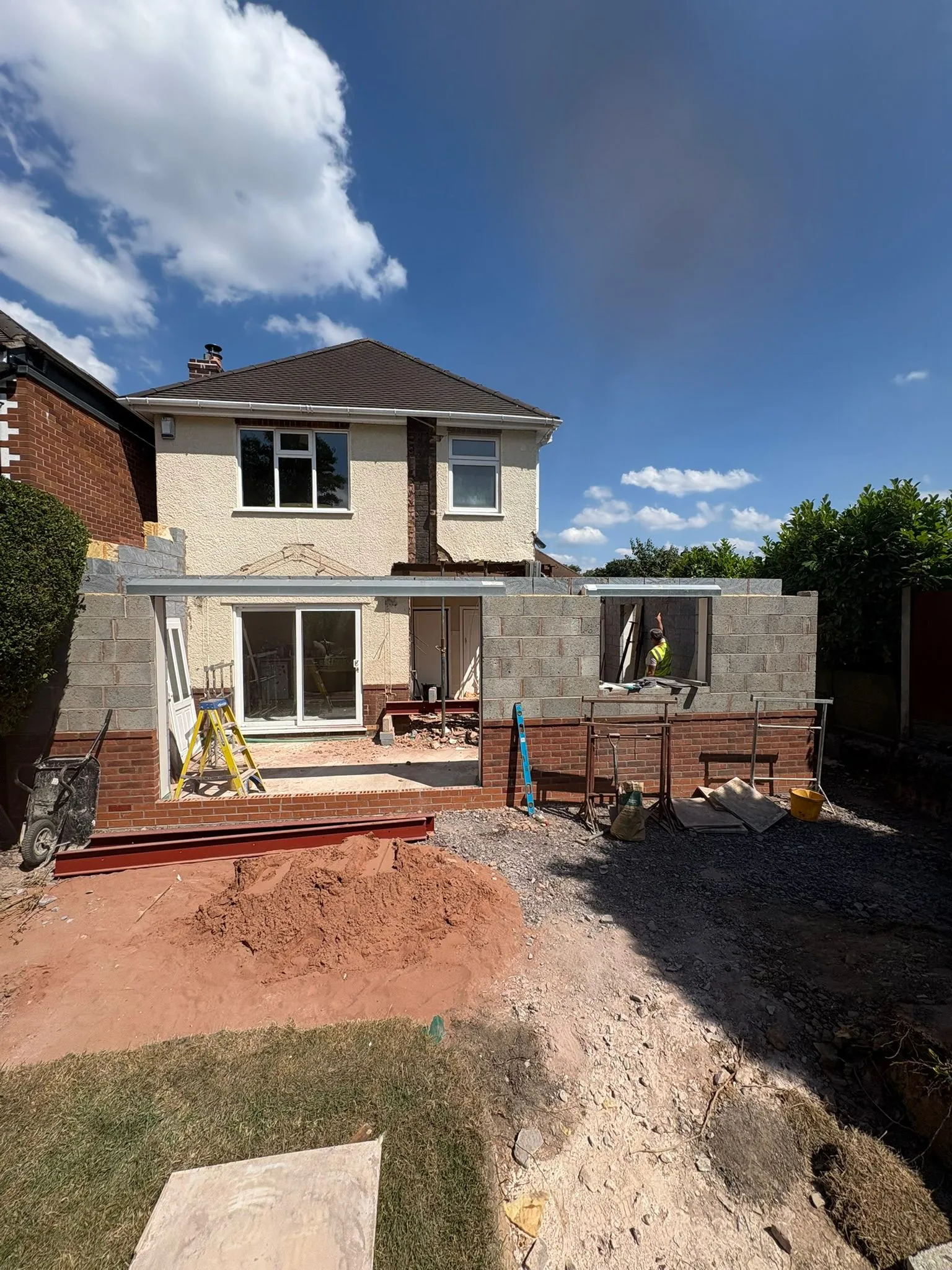 A house with a single-storey home extension in the early stages of construction, featuring new brickwork, concrete block walls, and exposed steel beams, with a builder visible inside.