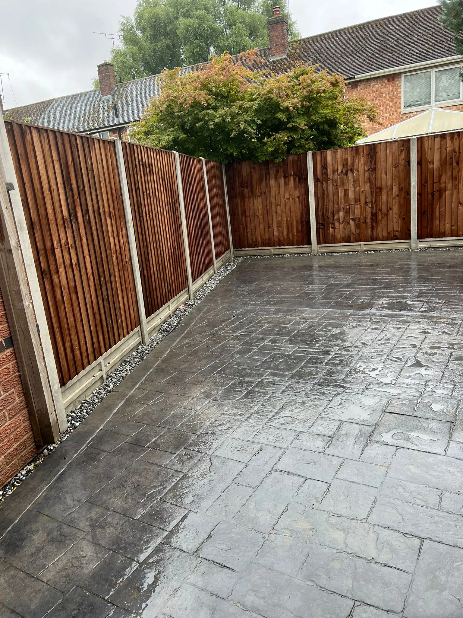 Newly laid patterned concrete paving and dark timber closeboard fencing with concrete posts and gravel boards, wet after installation or rain, showing a complete garden transformation.