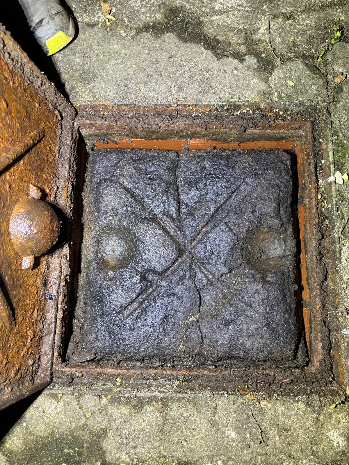 A photo showing a severely blocked outdoor drain or manhole filled with dark sludge, with a worker's boot visible nearby.