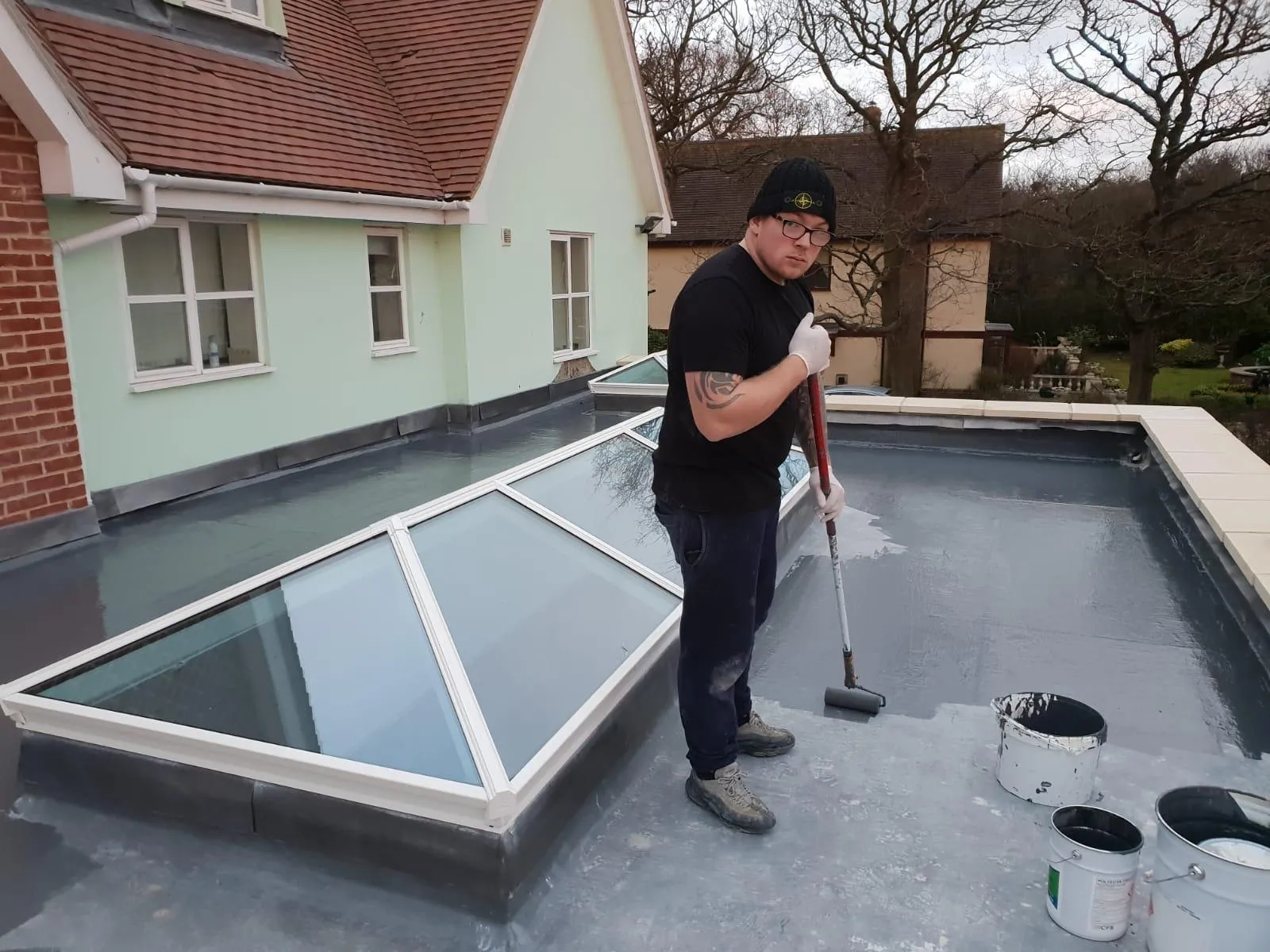 A tradesperson applying a dark grey GRP fibreglass coating to a new flat roof with large rooflights/lanterns, using a roller, with paint buckets visible.