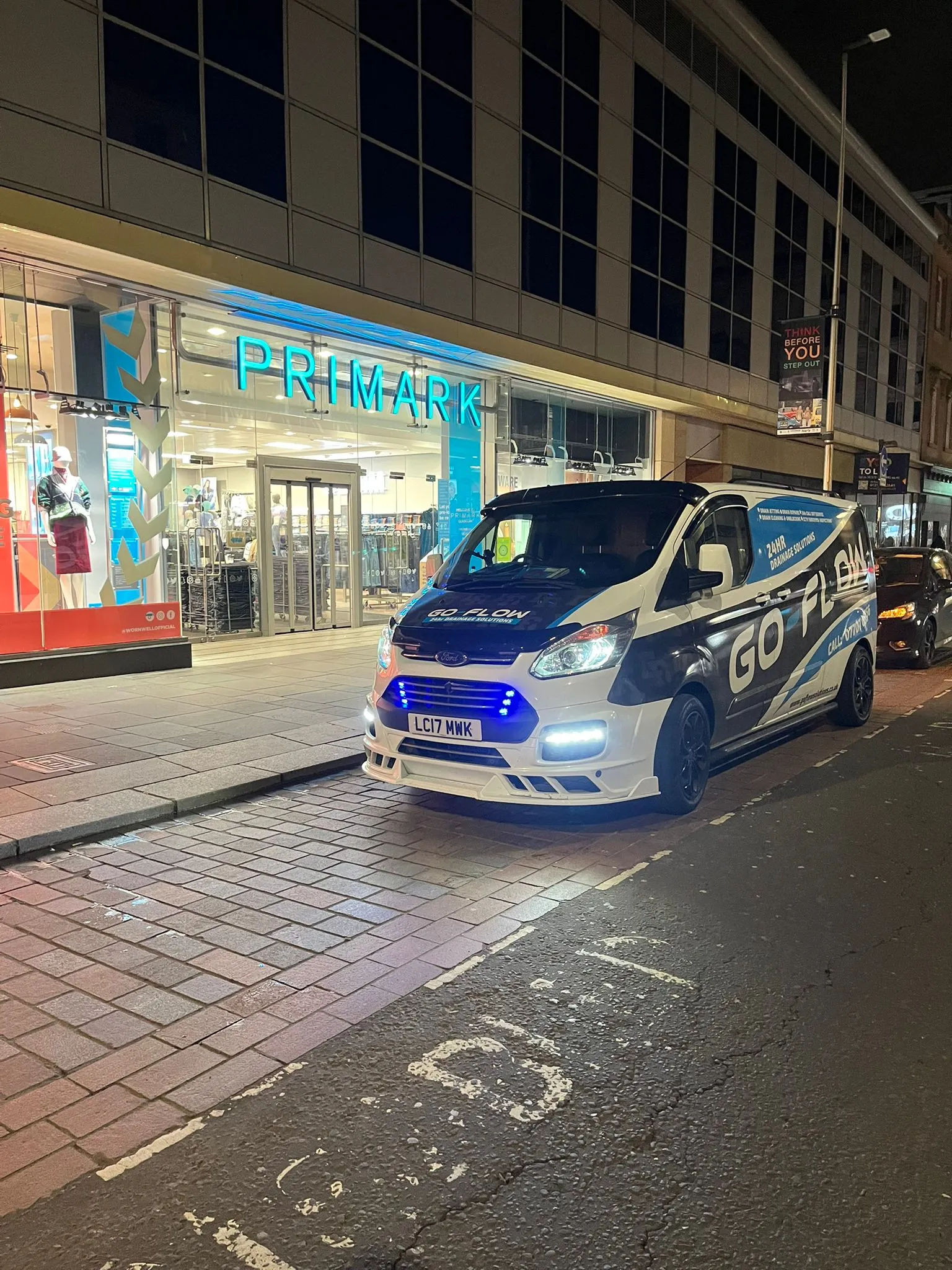 A white and blue Go Flow Solutions Edinburgh branded service van with its lights on, parked on a cobbled street in front of a Primark shop at night. The van advertises 24-hour drainage solutions.