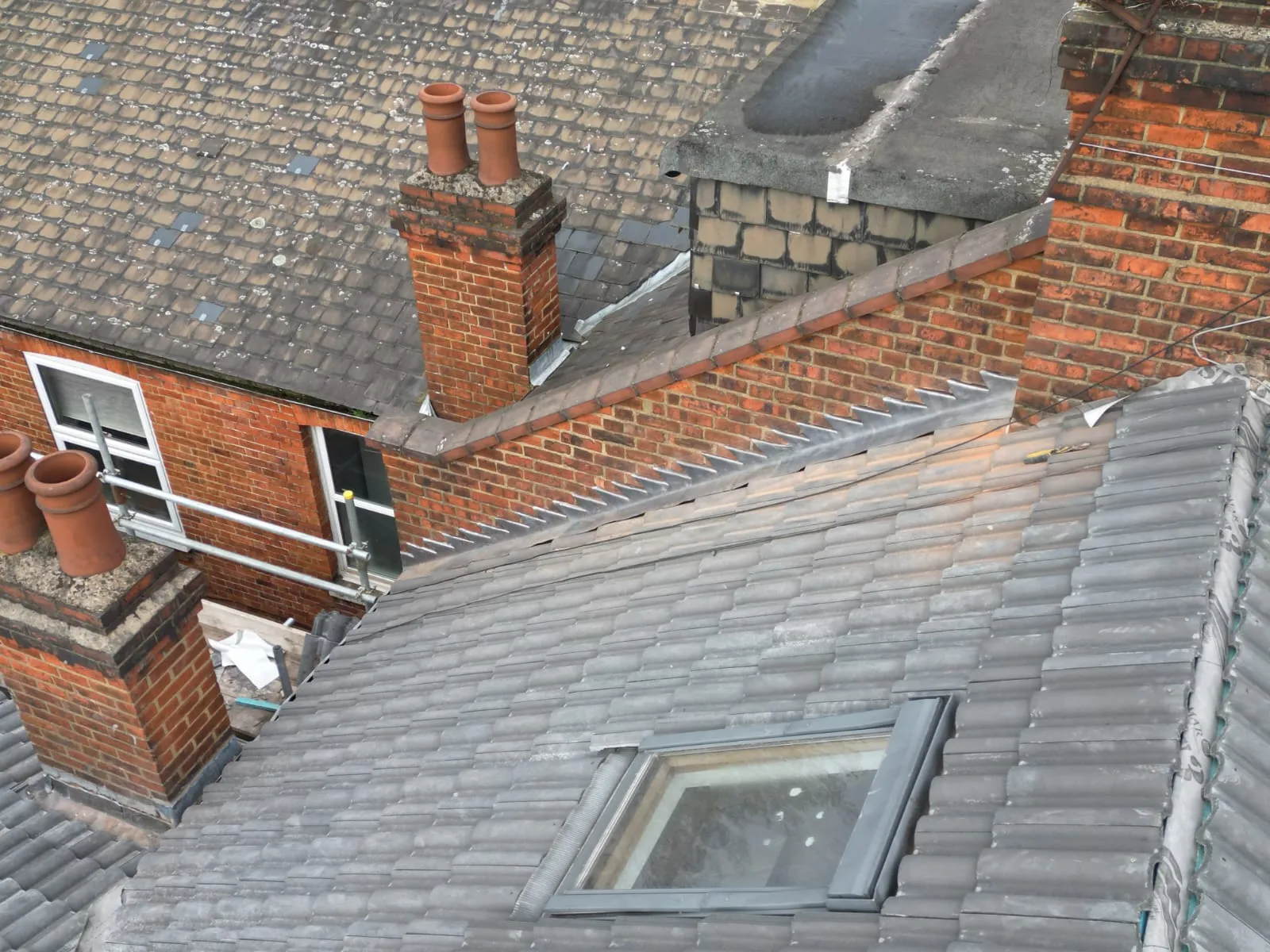 Aerial view of residential roofs with tiled pitches, a flat roof, chimneys, a skylight, and scaffolding.