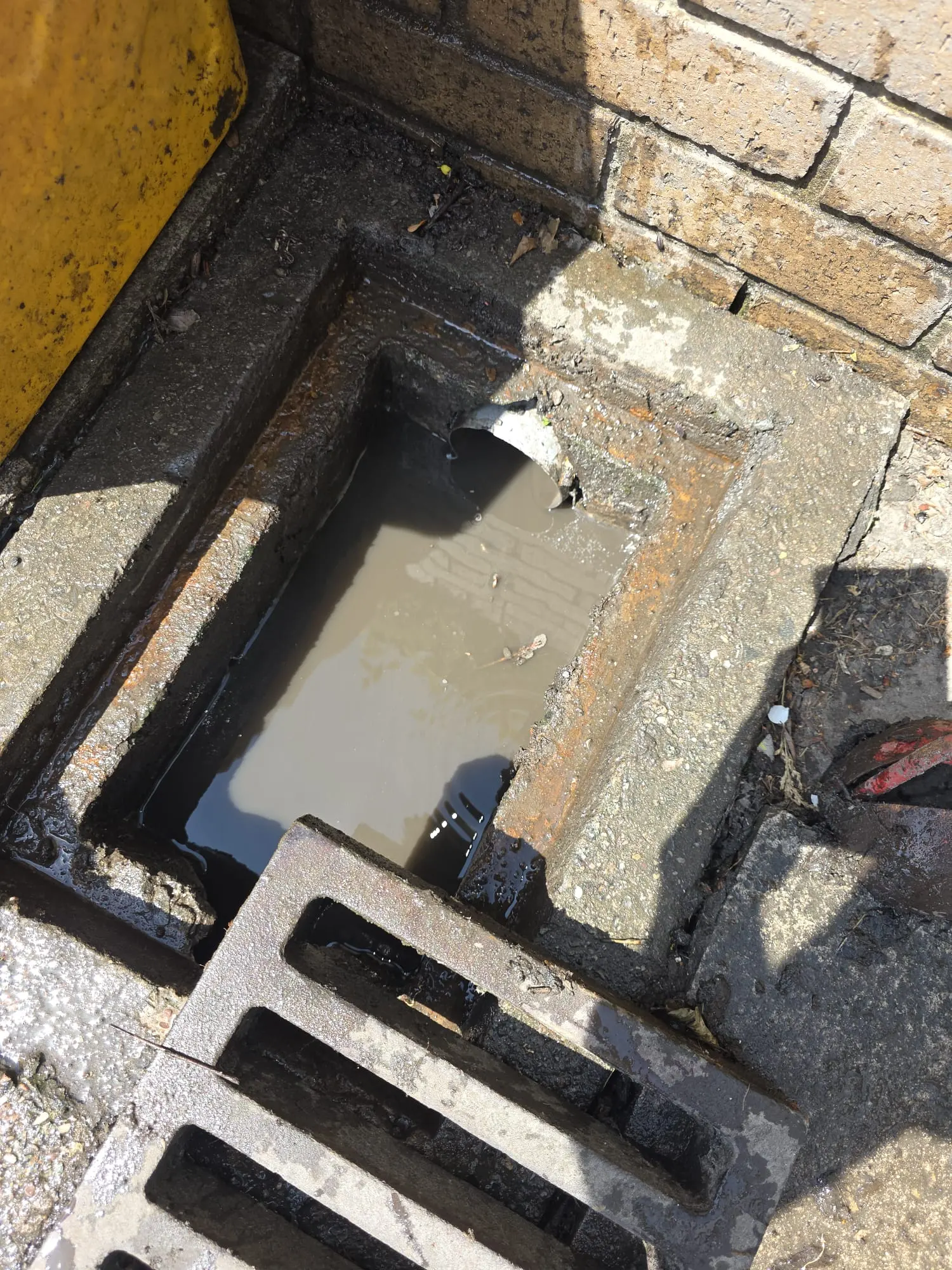 An open, blocked exterior drain filled with murky water next to a brick wall