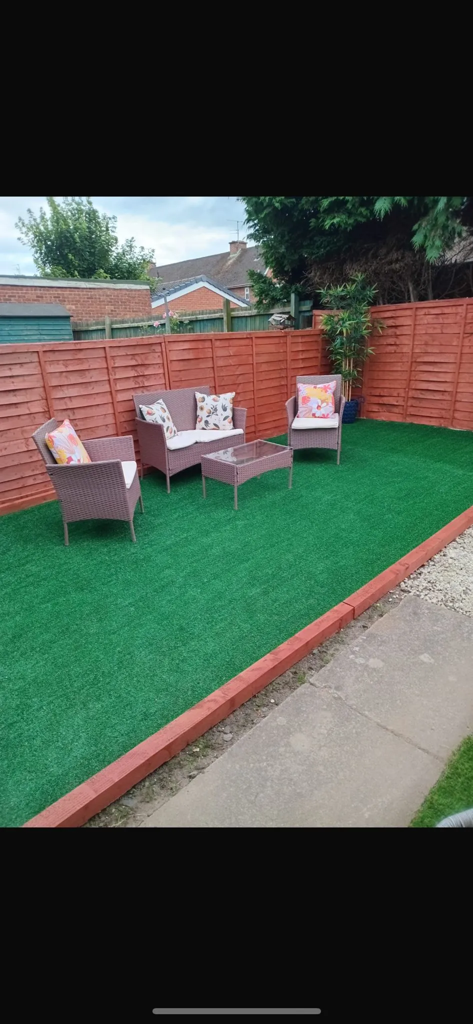 A modern, low-maintenance garden with bright green artificial grass, a grey rattan sofa, two matching chairs, and a glass-topped coffee table, all set against recently installed red-stained wooden fence panels. A potted bamboo plant stands in the corner.