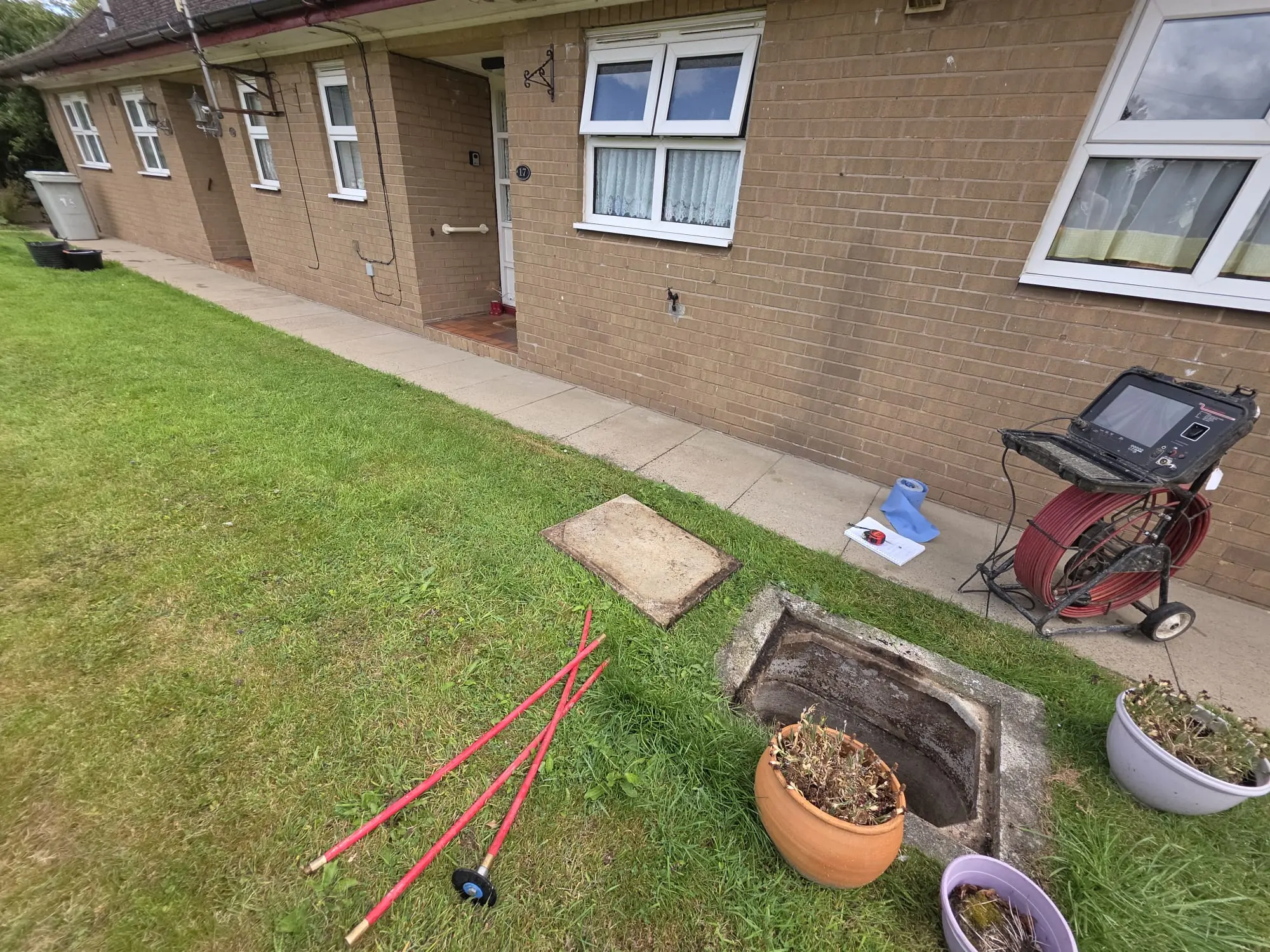CCTV drain survey camera and red drain rods next to an open manhole on a residential lawn