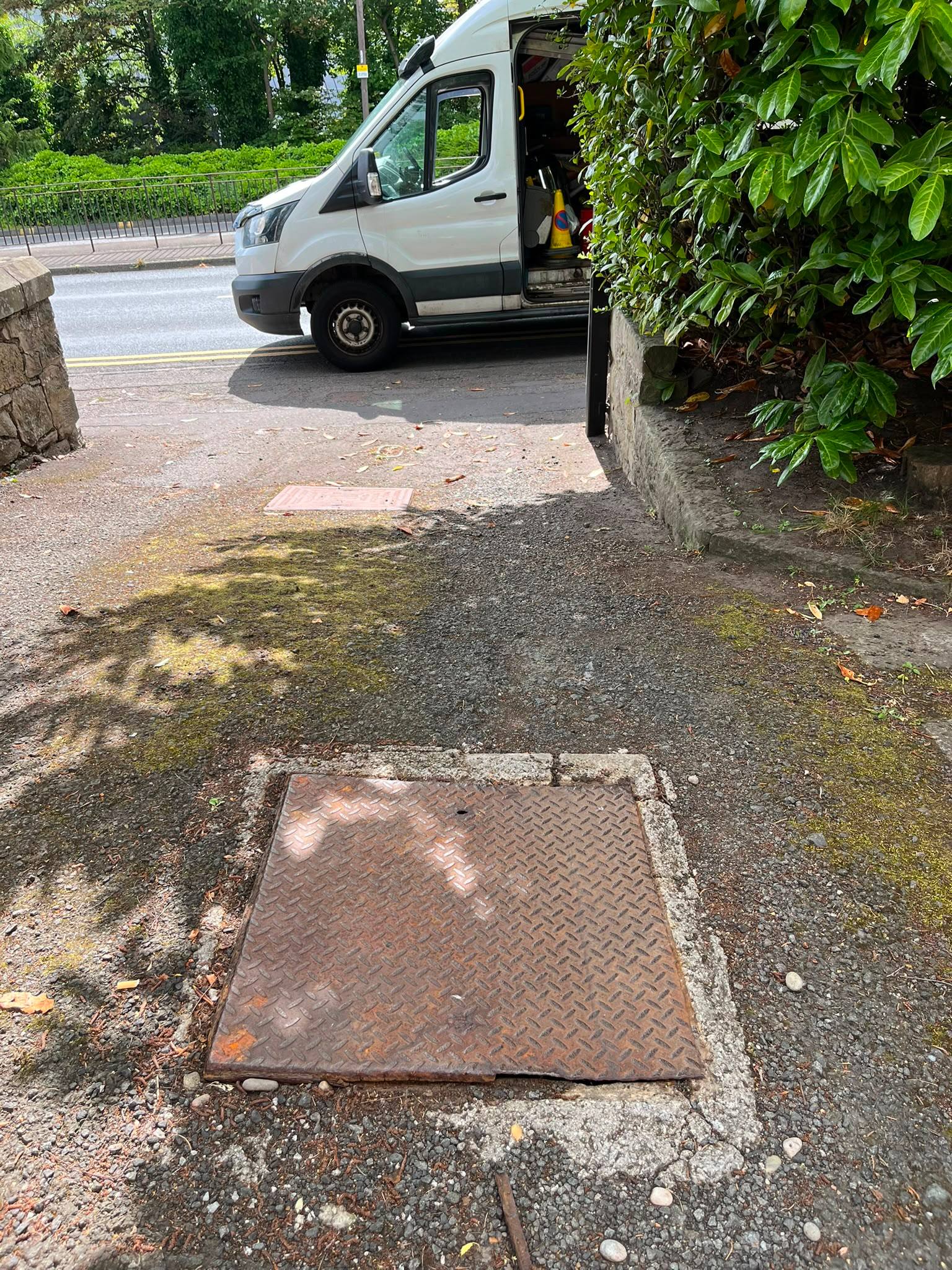 A rusty manhole cover on a residential pathway with a white CB Drainage van parked on the street, preparing for professional manhole cleaning and drain inspection.