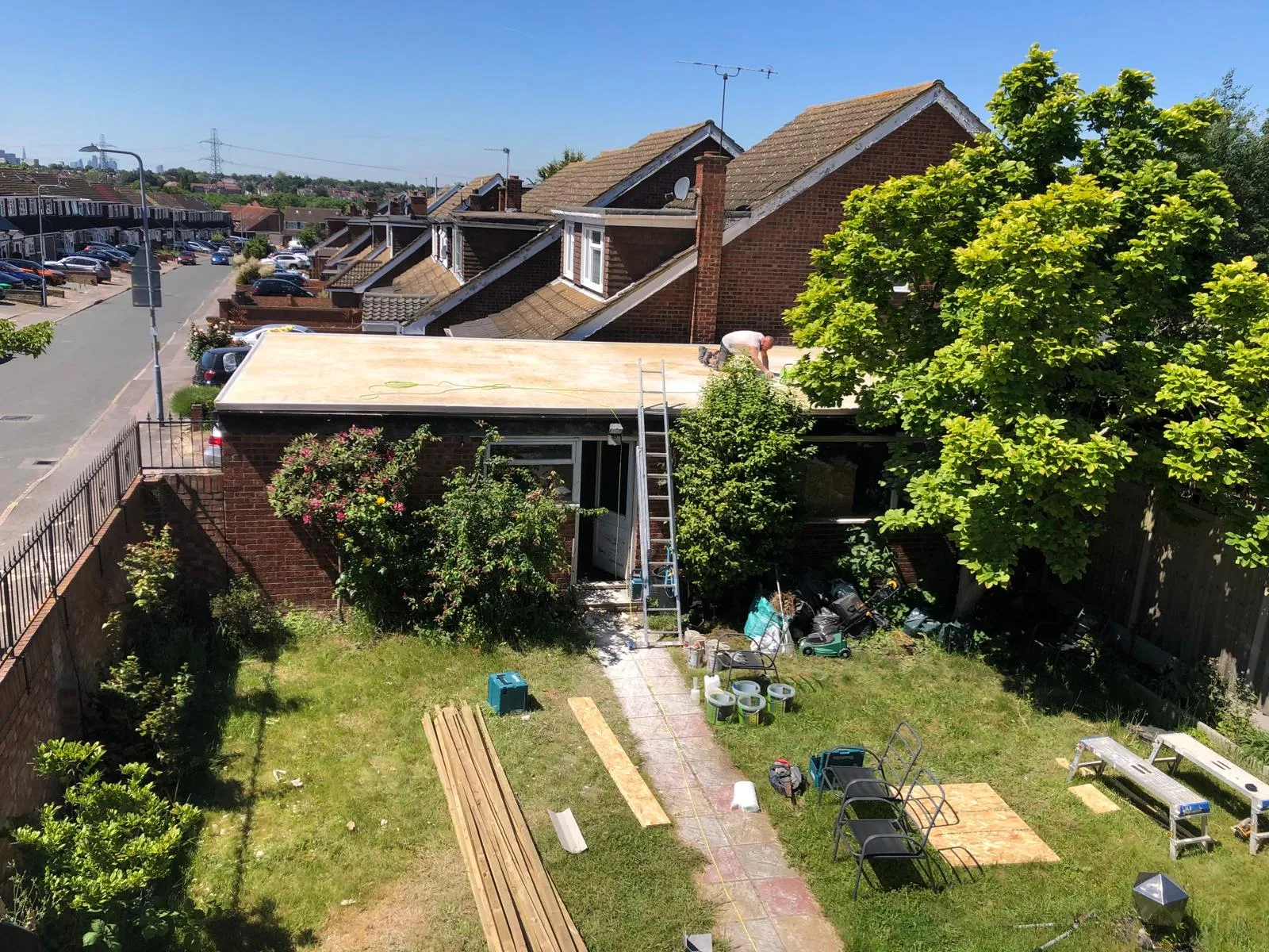 Builder working on a new flat roof installation or repair on a house extension, with a ladder, timber, and other construction materials in the garden.