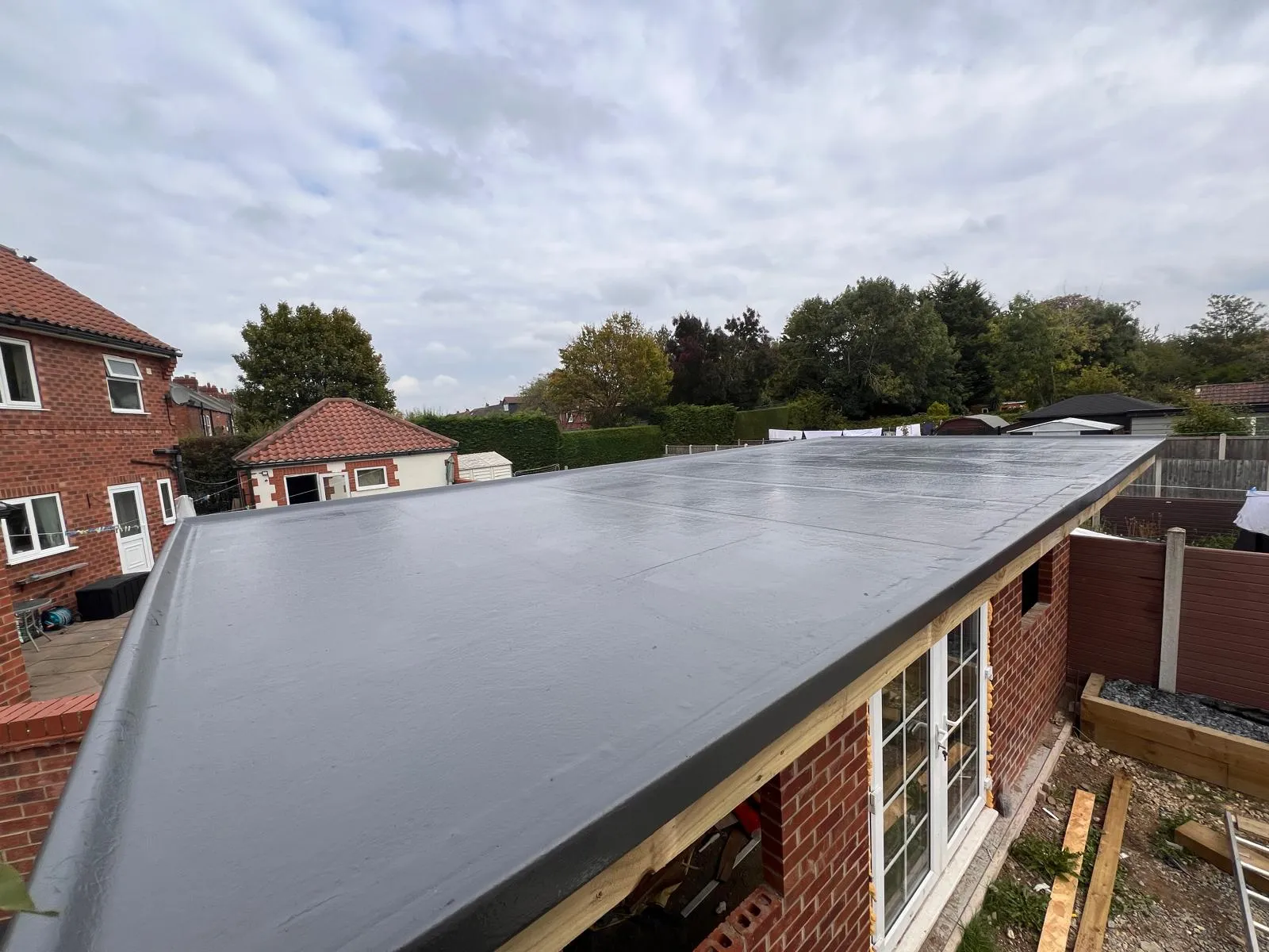 A close-up view of a newly installed dark grey GRP fibreglass flat roof on a building extension, with residential properties and trees in the background under a cloudy sky.