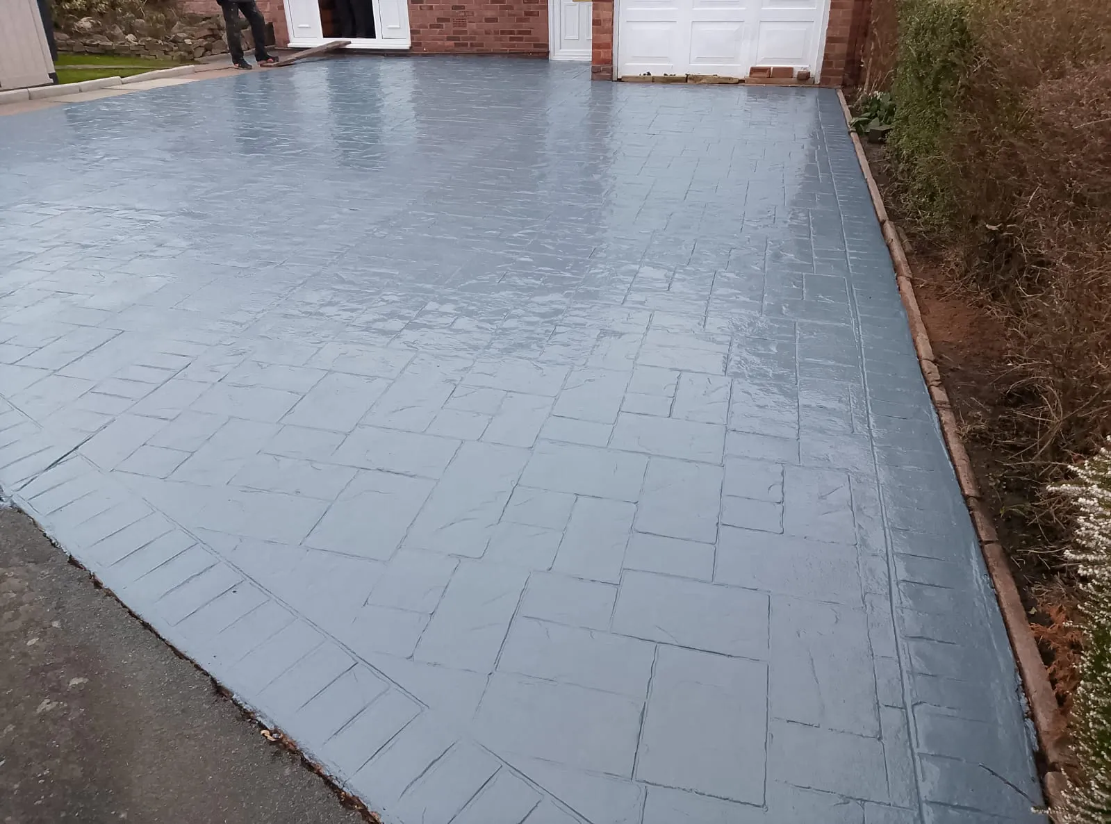 A new, grey pattern imprinted concrete driveway with a shiny, sealed finish leading up to a house with a white garage door and front entrance.
