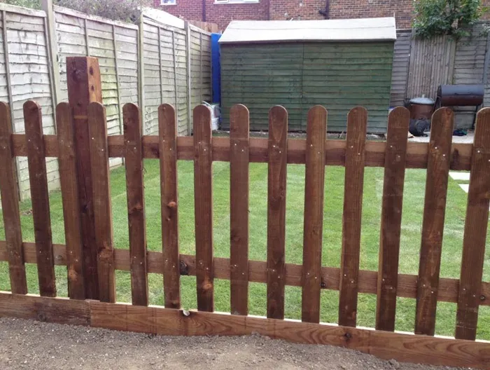 A newly installed dark brown wooden picket fence in a garden, with a green lawn and shed in the background.