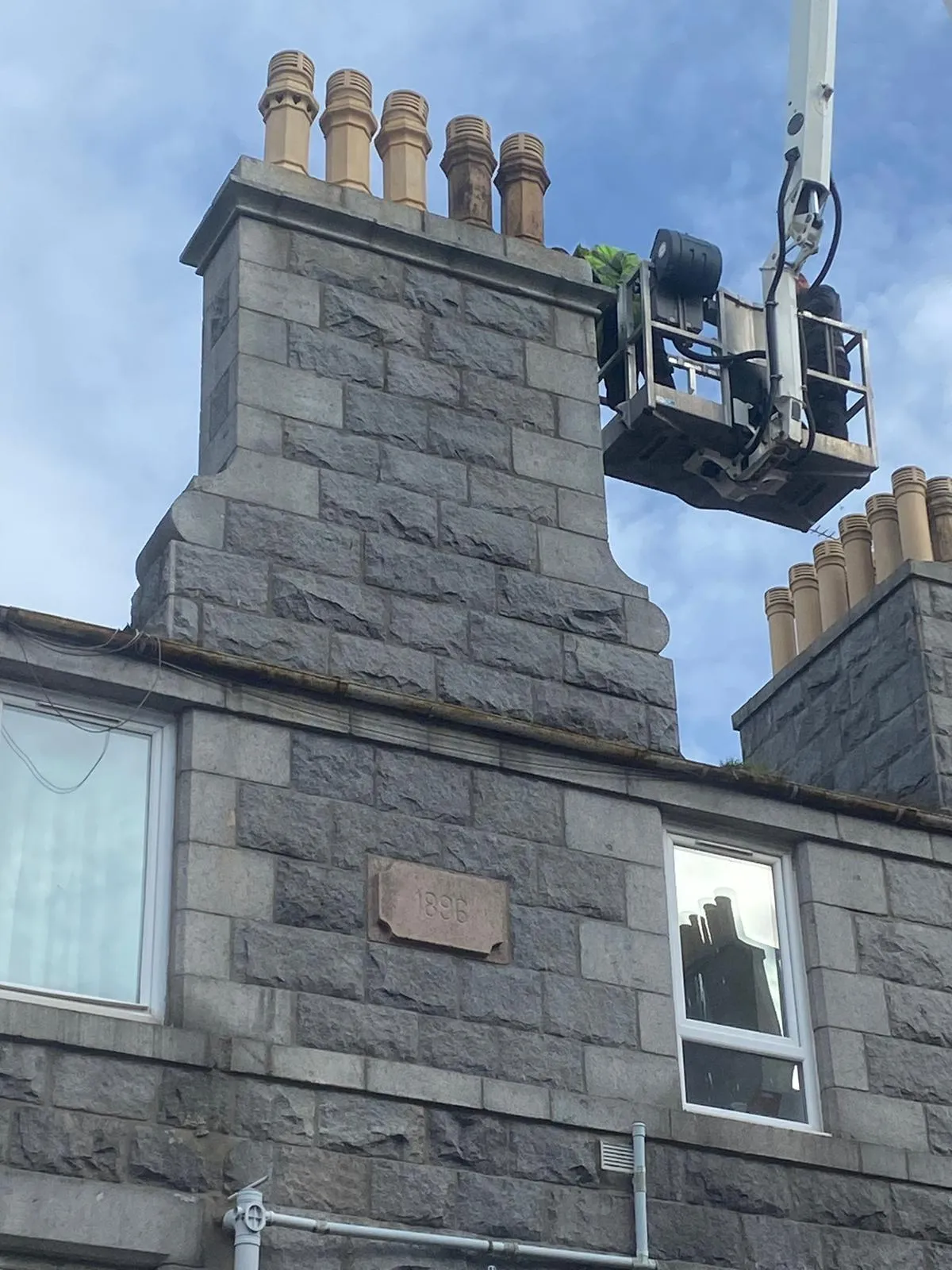 A worker in a cherry picker repairing or maintaining a tall, grey stone chimney stack with several chimney pots on an old building.