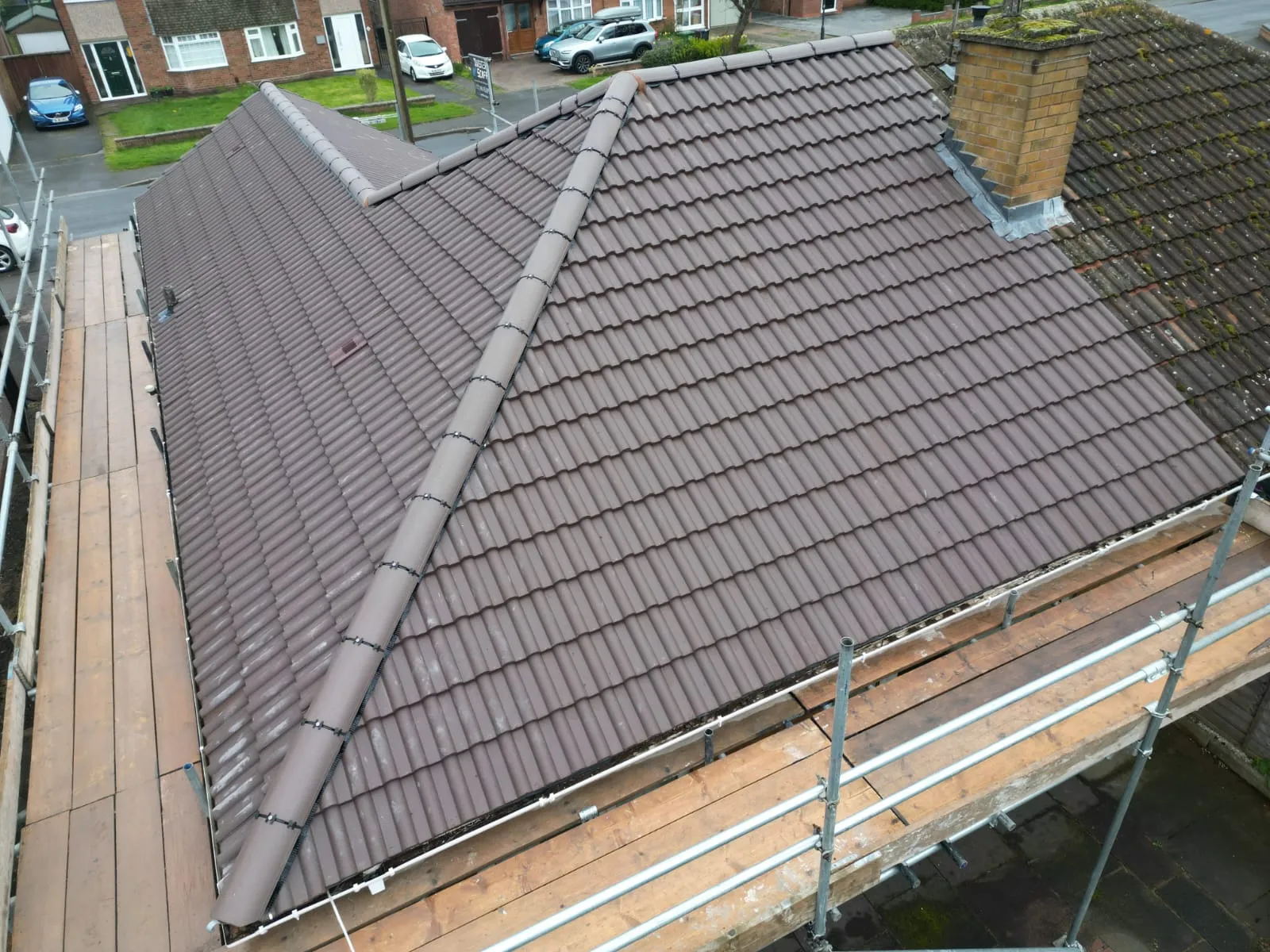 Newly installed or cleaned brown tiles on a pitched roof next to an old, mossy section and a brick chimney, with scaffolding, indicating roofing renovation.