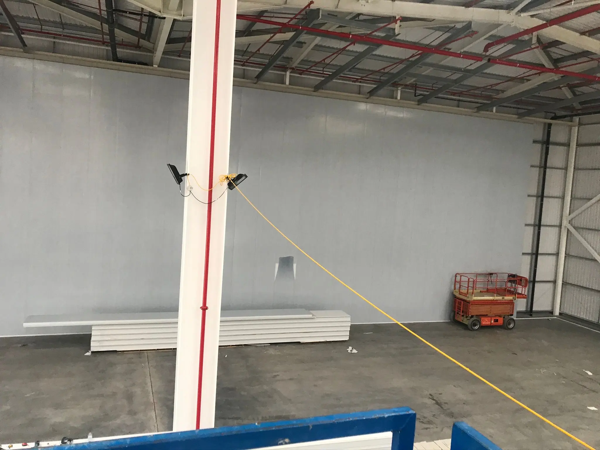 Interior of a newly constructed steel-framed industrial warehouse showing installed wall cladding, a steel support column, and a scissor lift on a concrete floor.