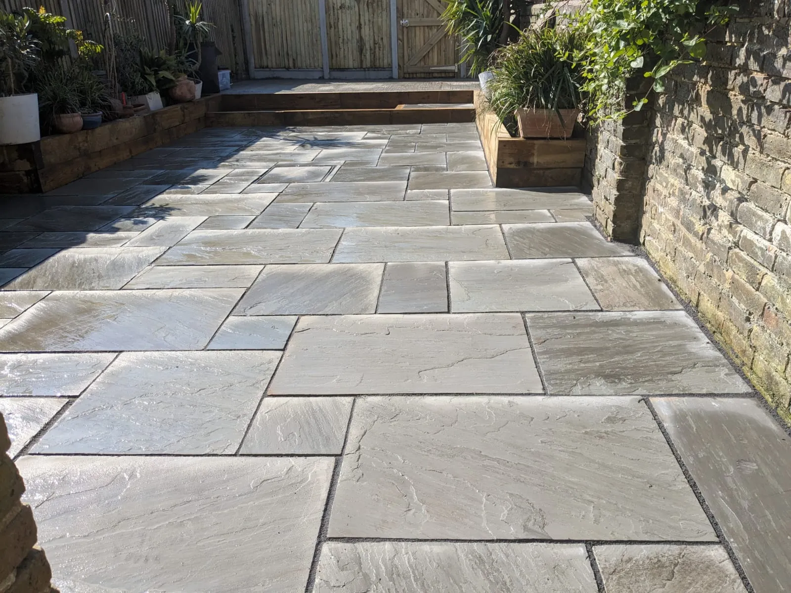 Newly laid Indian Sandstone patio with natural stone slabs, dark grout, wooden sleeper planters, and a wooden garden gate.