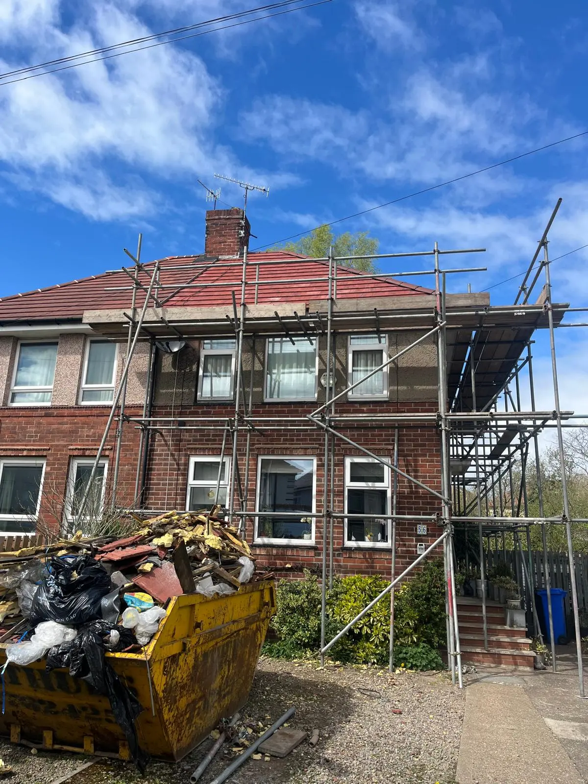 A residential roof replacement project in progress with scaffolding and a waste skip full of old tiles