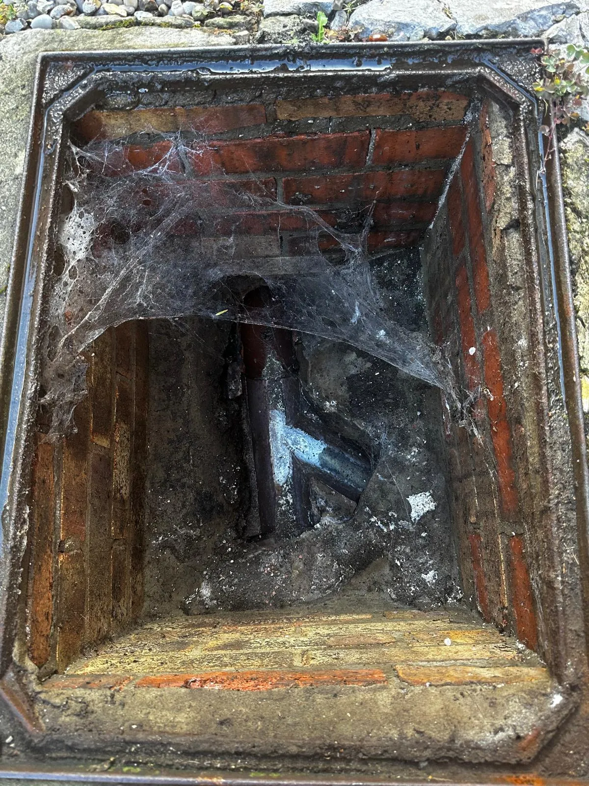 A residential drain inspection chamber or manhole, dirty and with cobwebs, used for accessing blocked or damaged drainage systems.