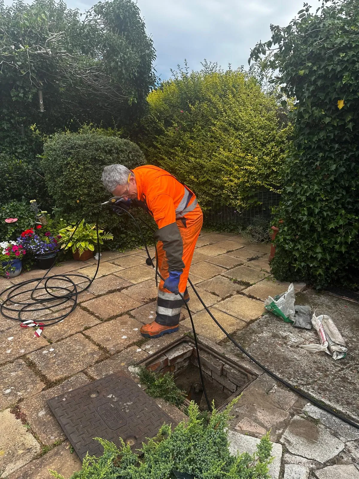 A drain unblocking technician examining an outdoor drain in a garden, with equipment for clearing blockages or conducting a CCTV survey.