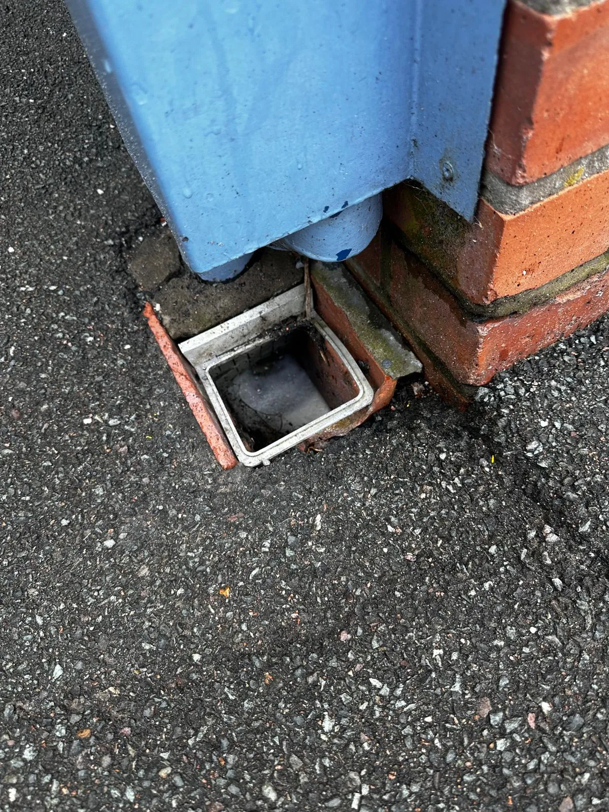 A close-up of an outdoor drainage gully next to a brick wall and tarmac, with a blue downpipe feeding into it and visible dirt suggesting a blockage or a need for cleaning.