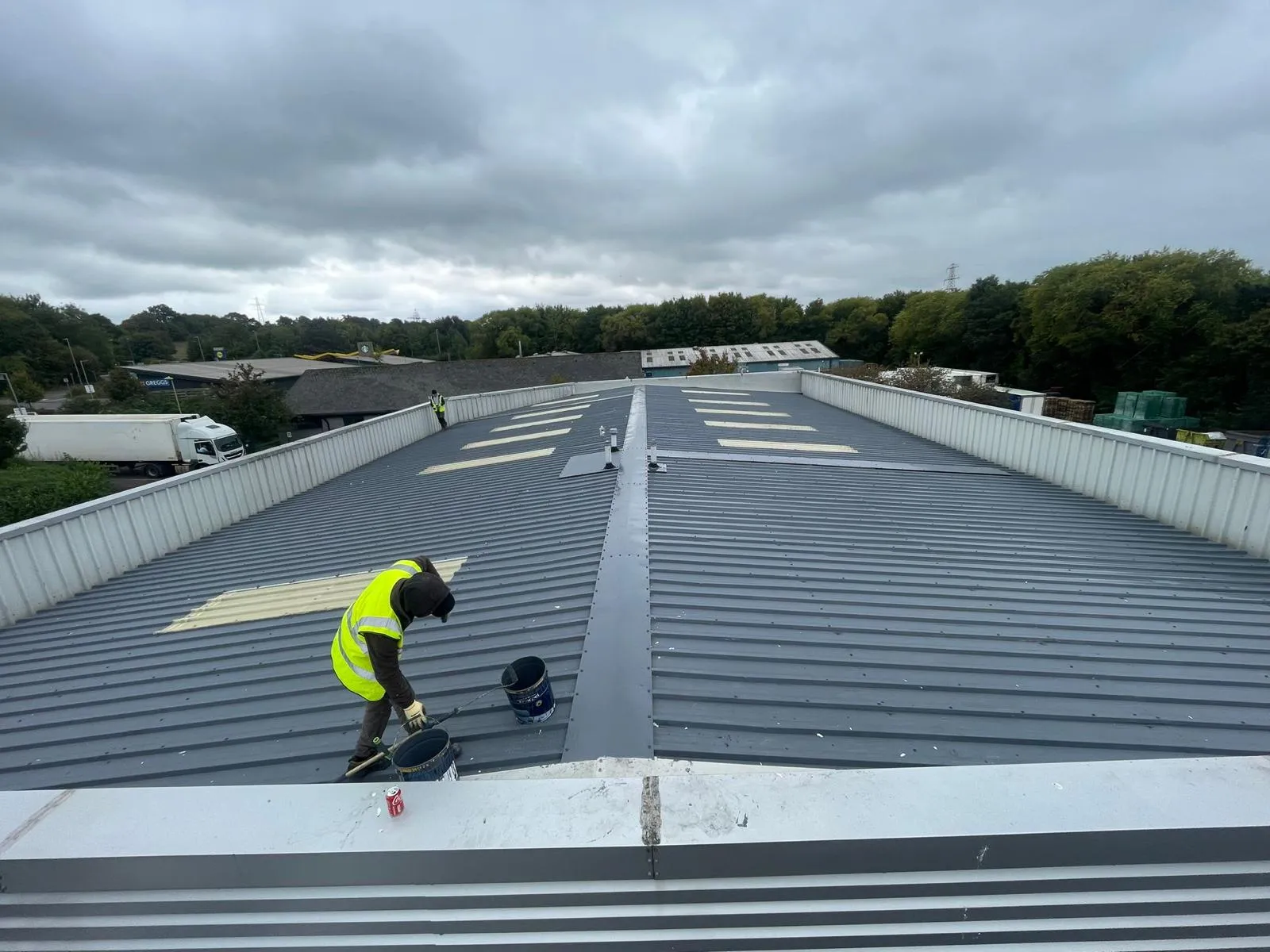 Two roofers working on a large commercial metal roof, one applying a coating to the surface, under an overcast sky. Visible in the background are trees, other buildings, and a white lorry.