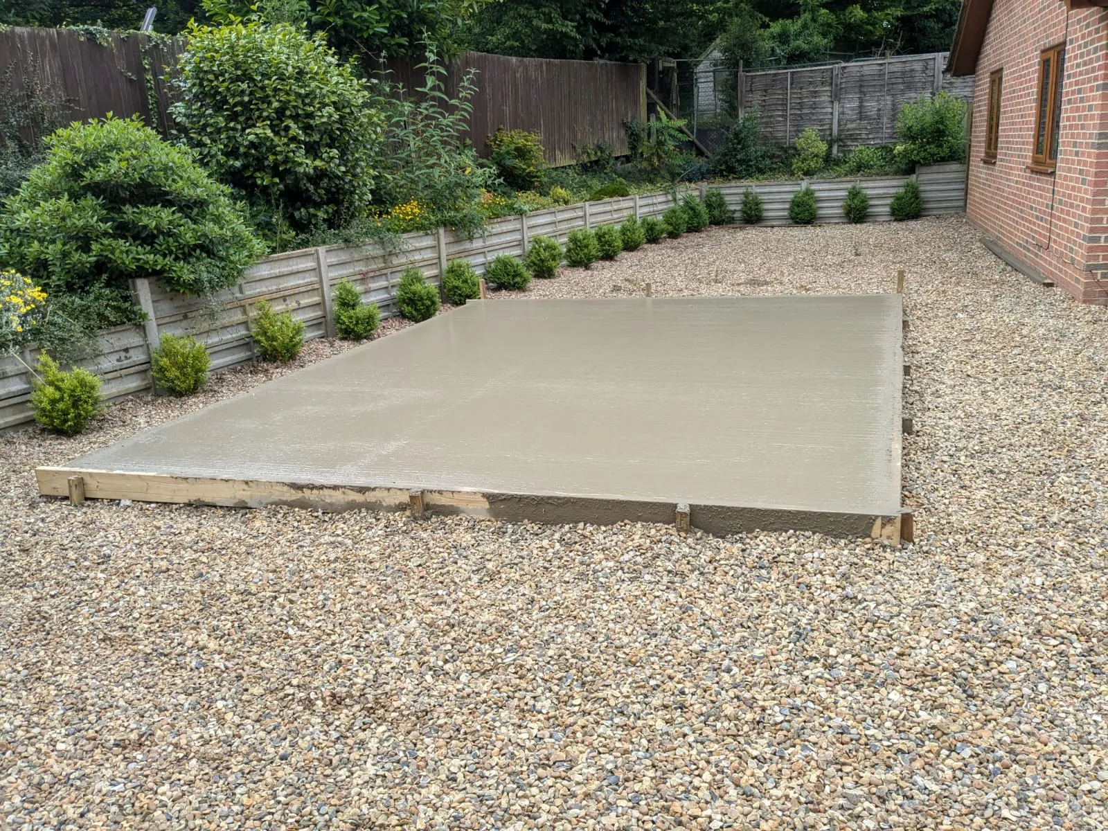 Newly poured concrete slab in a garden, surrounded by light gravel, with a fence and greenery in the background.