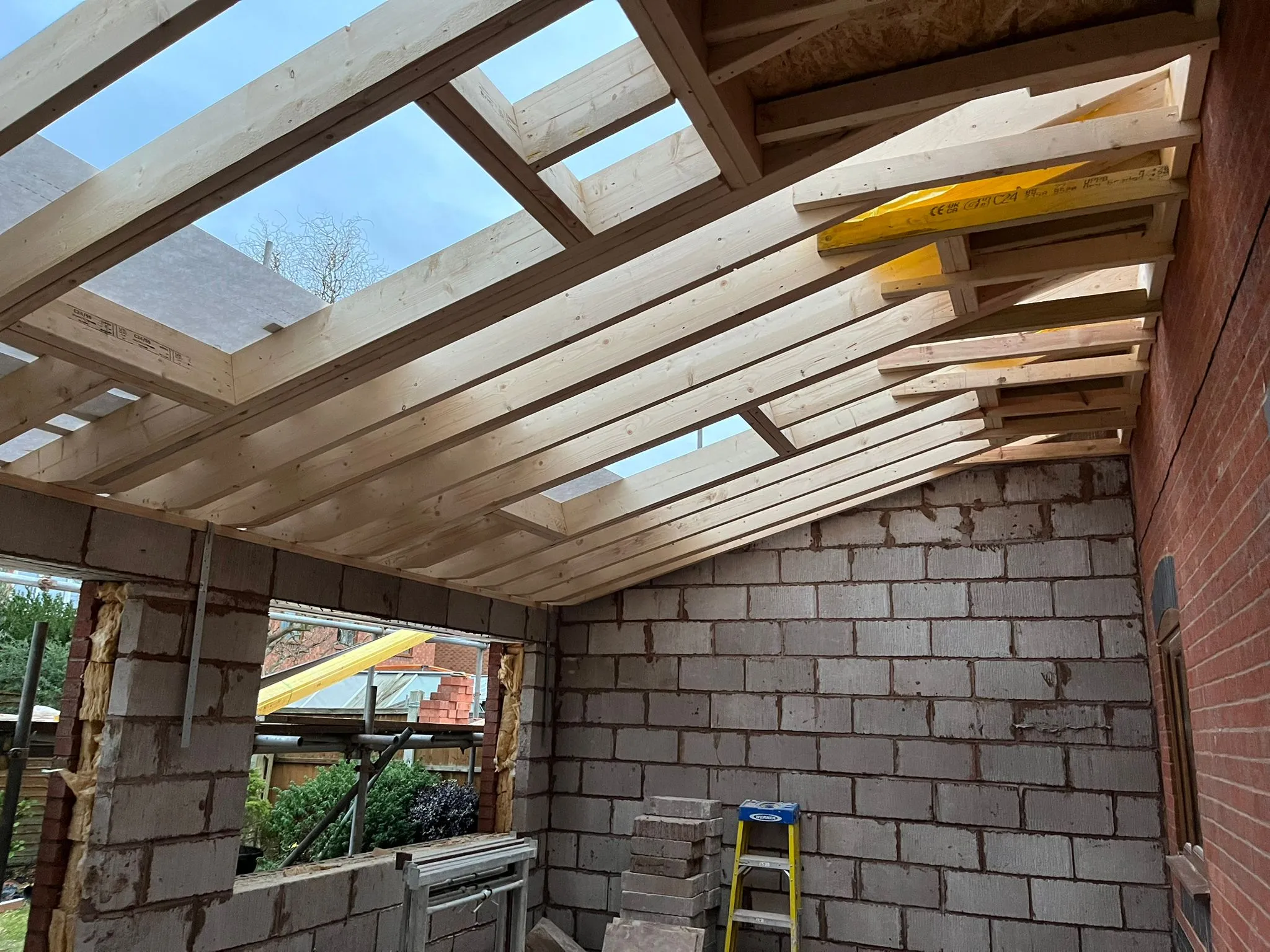 Interior view of a single-storey home extension during construction, highlighting the wooden roof structure with skylight openings, and the new blockwork walls alongside an existing brick wall.